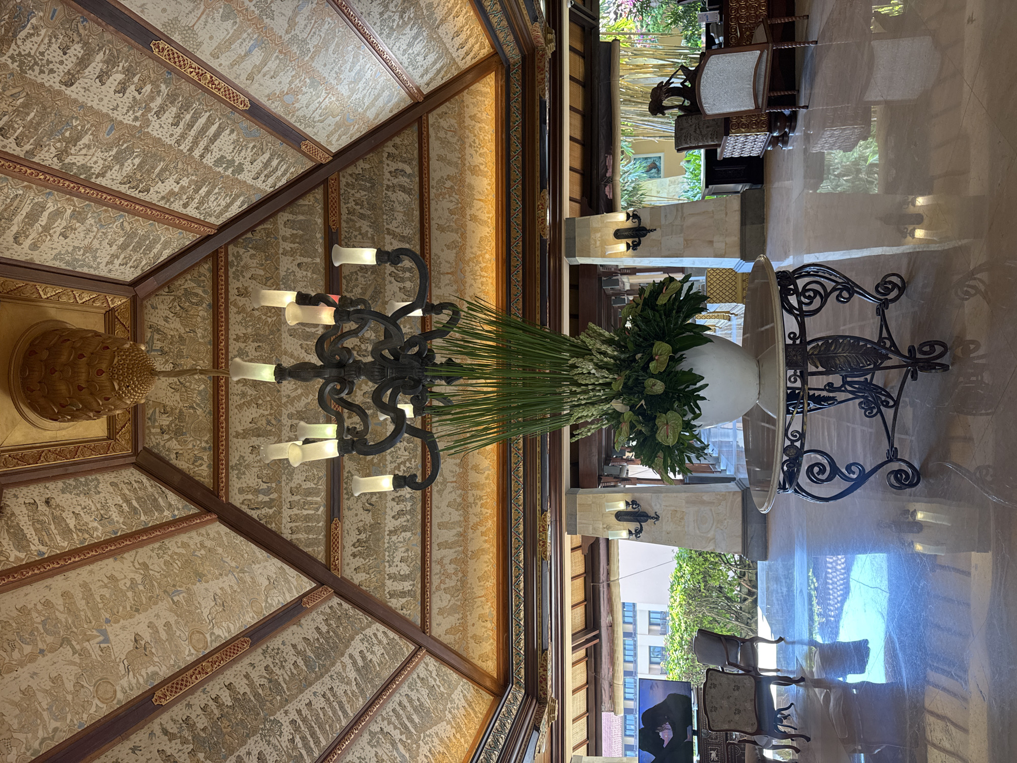 Indoor scene in Kuta Selatan, Indonesia. The view is upward to a coffered ceiling with intricate narrative artwork panels framed with dark wood. A black metal chandelier hangs from the ceiling. A large white spherical vase filled with green foliage stands on a black wrought-iron table. The highly polished floor reflects the scene and the outdoor environment visible through windows. Chairs are partially visible in the background.