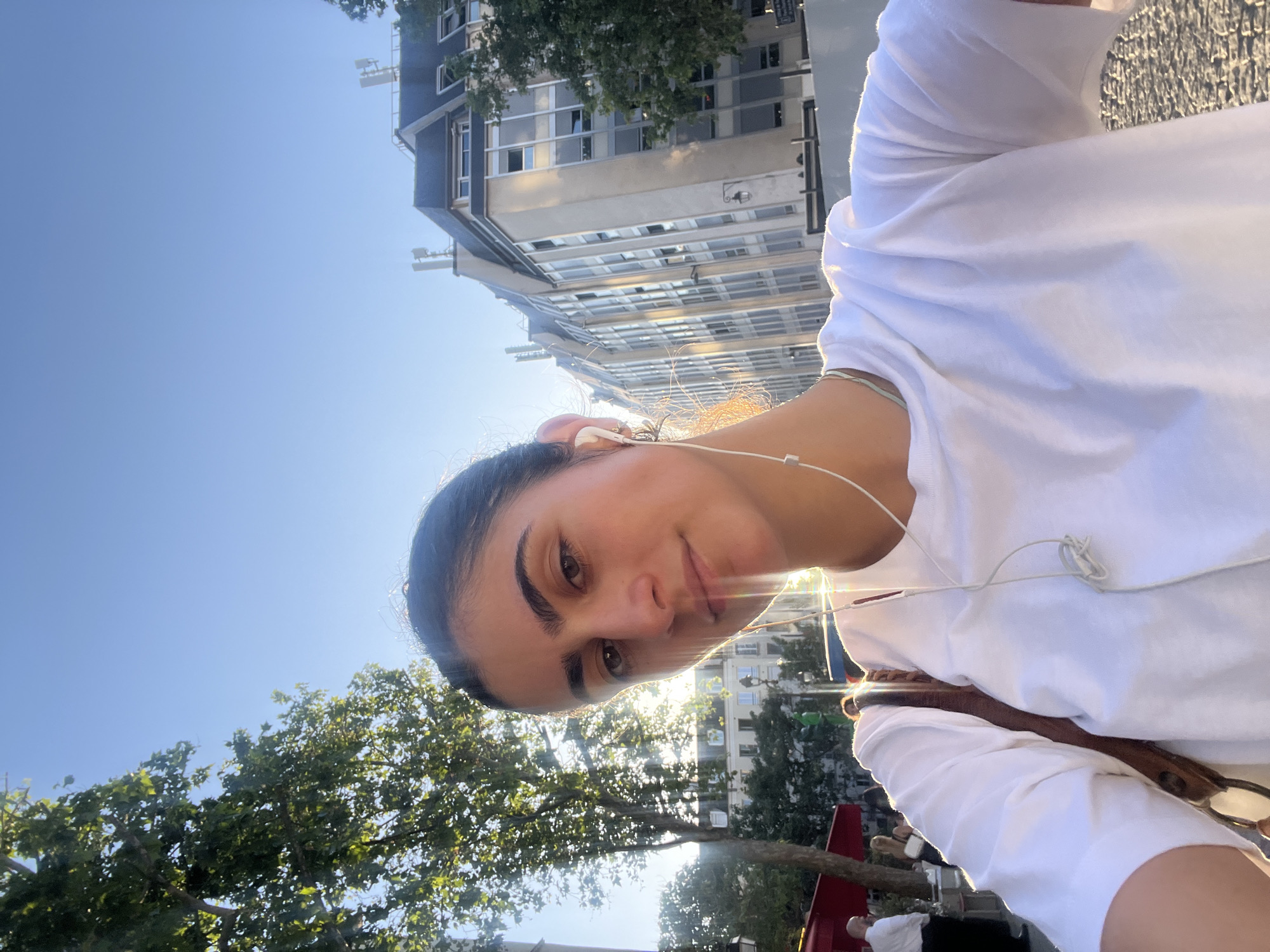 A daytime outdoor medium shot depicts a woman facing the camera with buildings and trees in the background. The woman has dark hair, brown eyes, and is wearing a white t-shirt and earphones. A brown bag strap is visible over her shoulder. The background includes trees, buildings with multiple windows, and a clear blue sky. The scene appears to be in Paris, France.