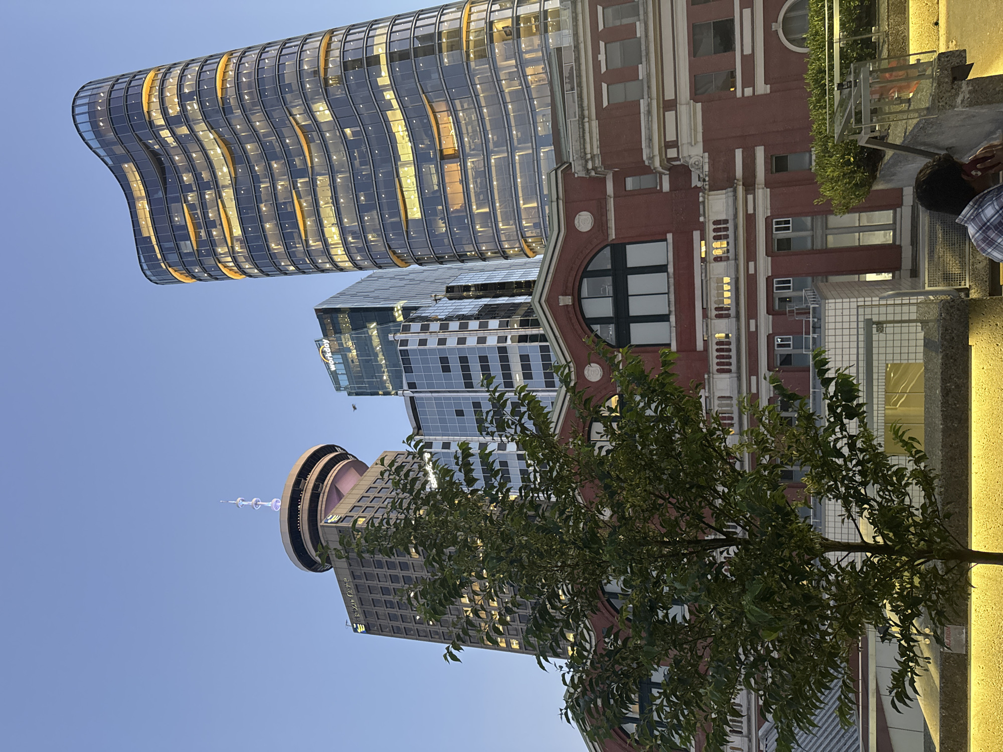 This image captures a view of the Vancouver Harbour Centre and surrounding buildings against a clear twilight sky. The Harbour Centre, a prominent circular building topped with a spire, is visible on the left, with the text "Harbour Centre" clearly displayed. To its right, a modern, wavy glass skyscraper with illuminated floors is the most striking feature, its curved facade reflecting the light. Adjacent to this is another glass skyscraper with a more rectangular design, bearing the "Workday" logo. In the foreground, a lower, red brick building with arched windows and classical architectural elements sits beneath the taller structures. A leafy green tree stands in front of the brick building, partially obscuring it. A person, wearing a plaid shirt, is seated on a concrete ledge in the lower right corner, appearing to be on a phone call. The scene is illuminated by artificial lights from the buildings and streetlights casting a warm glow on the concrete structures in the foreground.