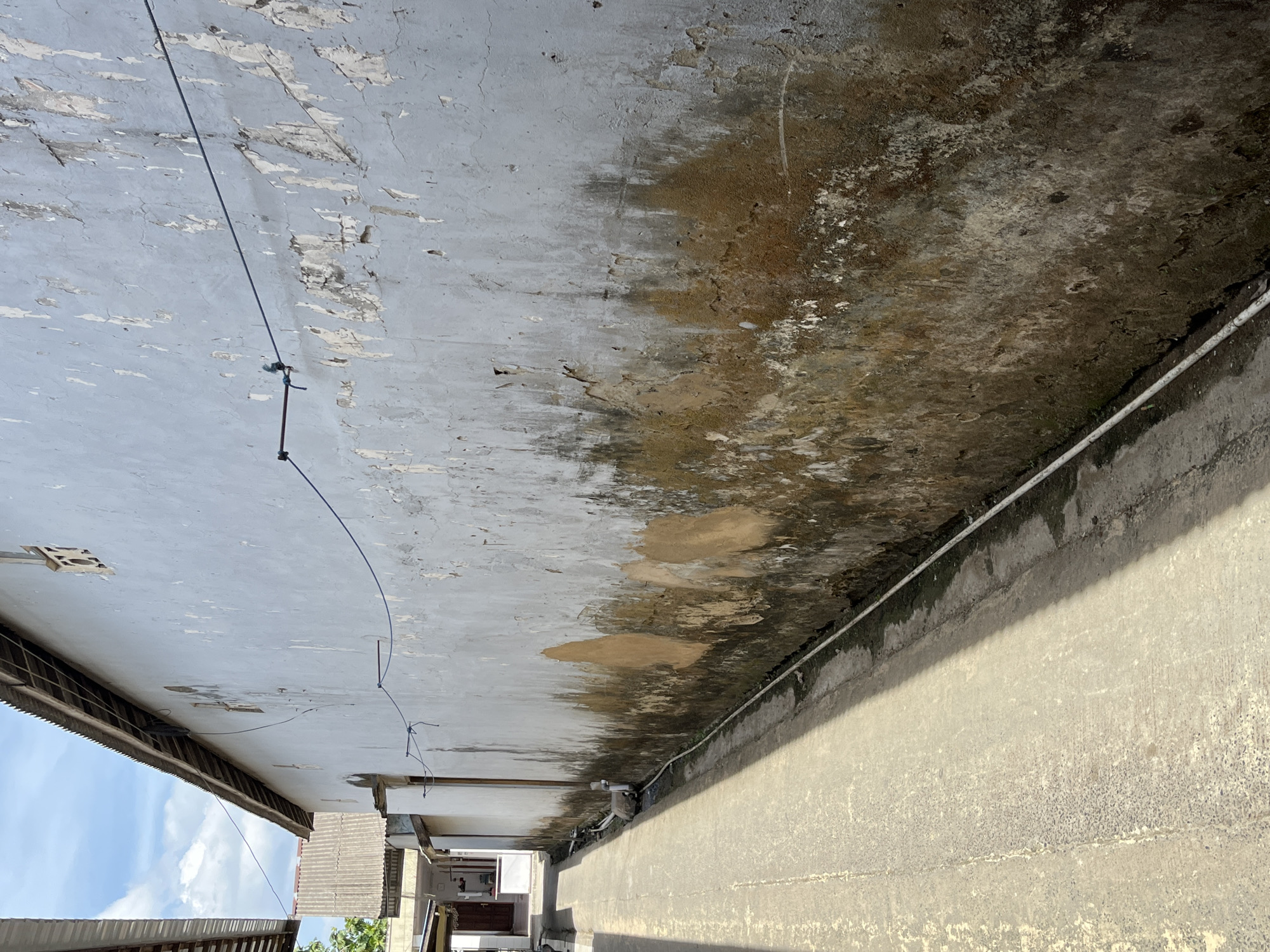 This is an exterior, eye-level, medium shot of a narrow alleyway with a tall, weathered wall on the right. The alleyway is paved with concrete and has a slight incline. The wall is painted a pale blue, but much of the paint is peeling, revealing patches of brown and grey plaster underneath. The lower portion of the wall is stained with dark, mossy growth. A white electrical conduit runs along the base of the wall. A thin blue wire is strung across the top of the wall, attached to rusty metal fixtures. In the distance, the alleyway opens slightly, revealing the side of a building with a corrugated metal roof. The sky above is blue with scattered white clouds. The scene appears to be in the city of Sejahtera, Indonesia.