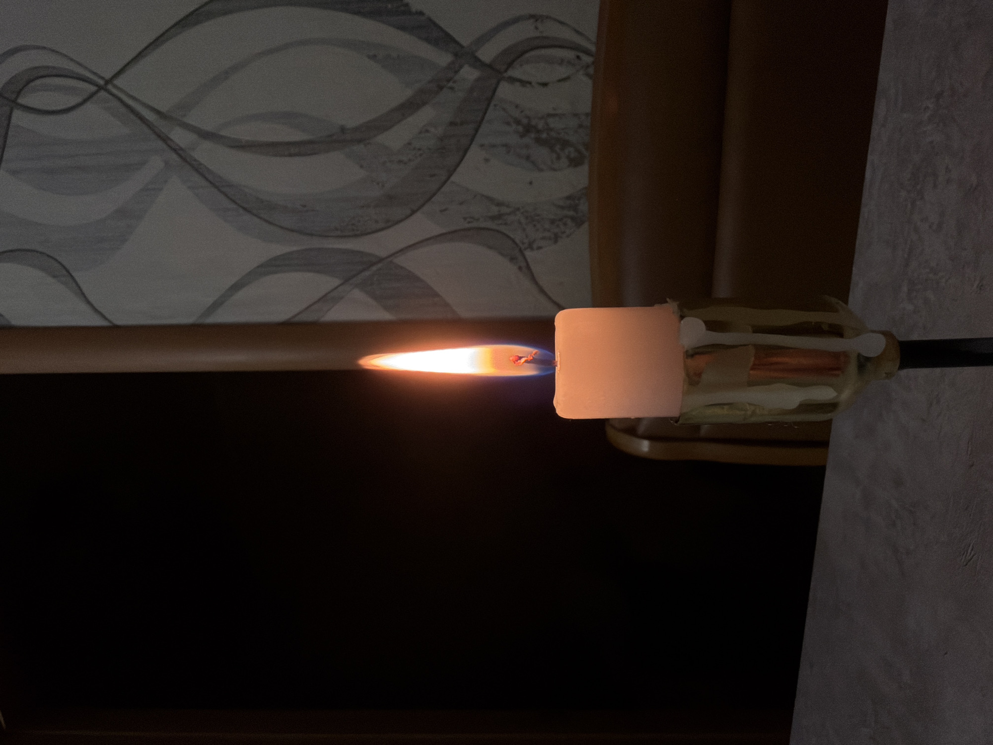 A lit candle is positioned horizontally in front of a dark background. Behind the candle are sections of a patterned wallpaper and what appears to be a portion of a brown couch. A glass-like or translucent container secures the base of the candle and contains cotton swabs and a thin rod. The location, Komsomolskiy, Russia, cannot be verified from the image.