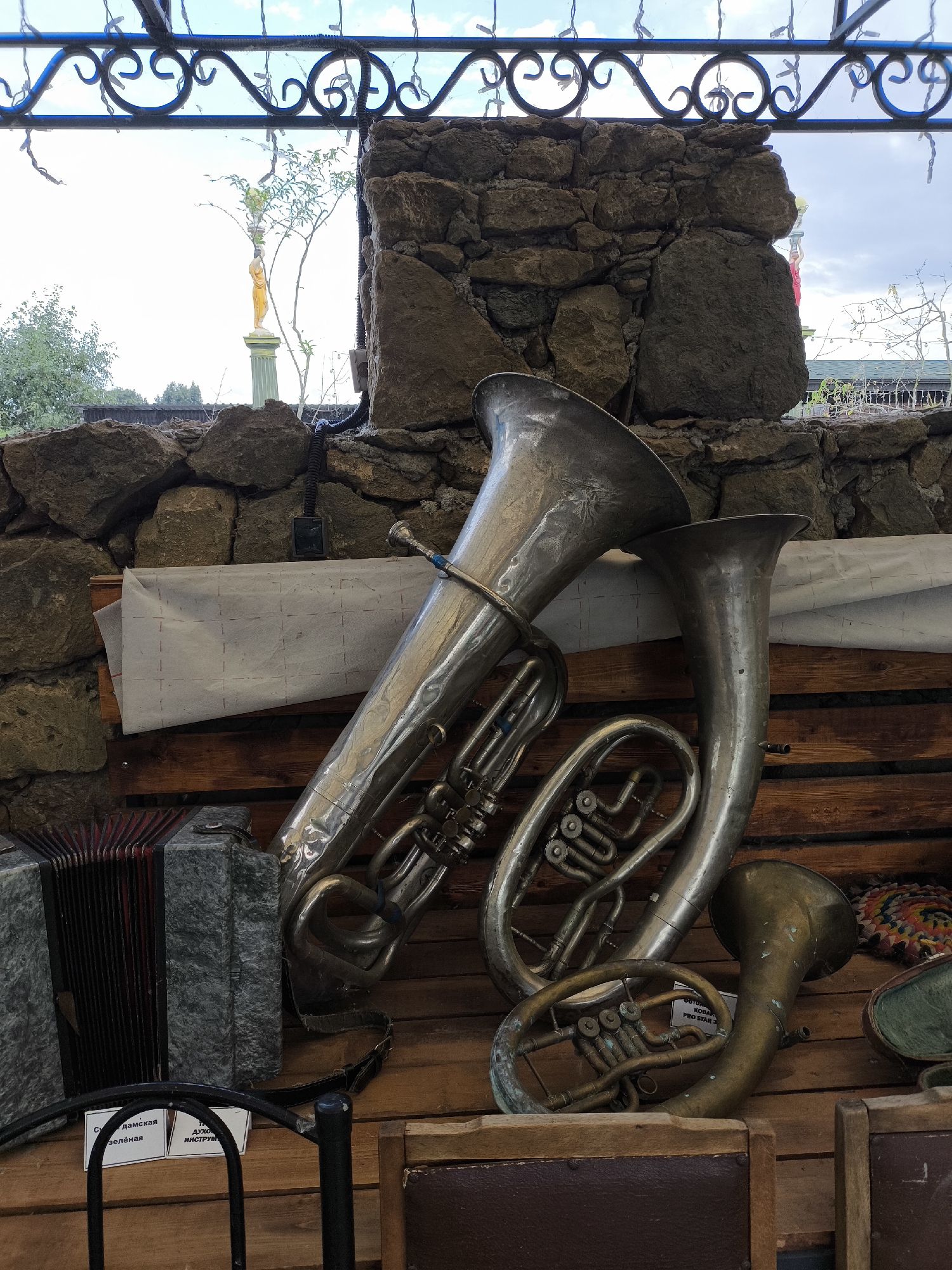 The image shows a collection of brass musical instruments, including a tuba, a euphonium, and a smaller horn, displayed on a wooden surface in front of a stone wall. To the left of the brass instruments is a grey accordion. A white cloth with a grid pattern is draped over wooden planks behind the instruments. Above, a metal railing with decorative curls runs along what appears to be an open-air structure, with a bright sky visible. In the distance, through an opening in the stone wall, a small yellow statue on a green pedestal can be seen, along with trees and some buildings. The overall setting suggests an outdoor display, possibly in a museum or a cultural space, located in Borgustanskaya, Russia. There are no people visible in the image.