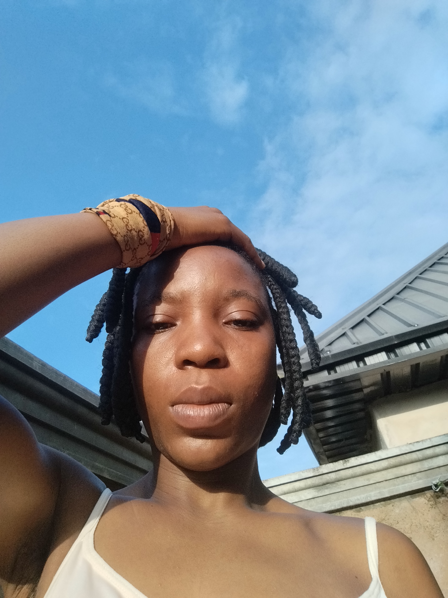 The image is a low-angle, close-up selfie of a Black individual with dark braided hair, wearing a white tank top and a patterned headband. The person's right hand is raised, resting on their head. The background consists of a blue sky with some clouds and the roofline of a building, suggesting an outdoor urban setting in Isiehor, Nigeria. The lighting suggests it is daytime, with the sun casting shadows on the individual's face and skin. The overall impression is a casual, outdoor portrait.