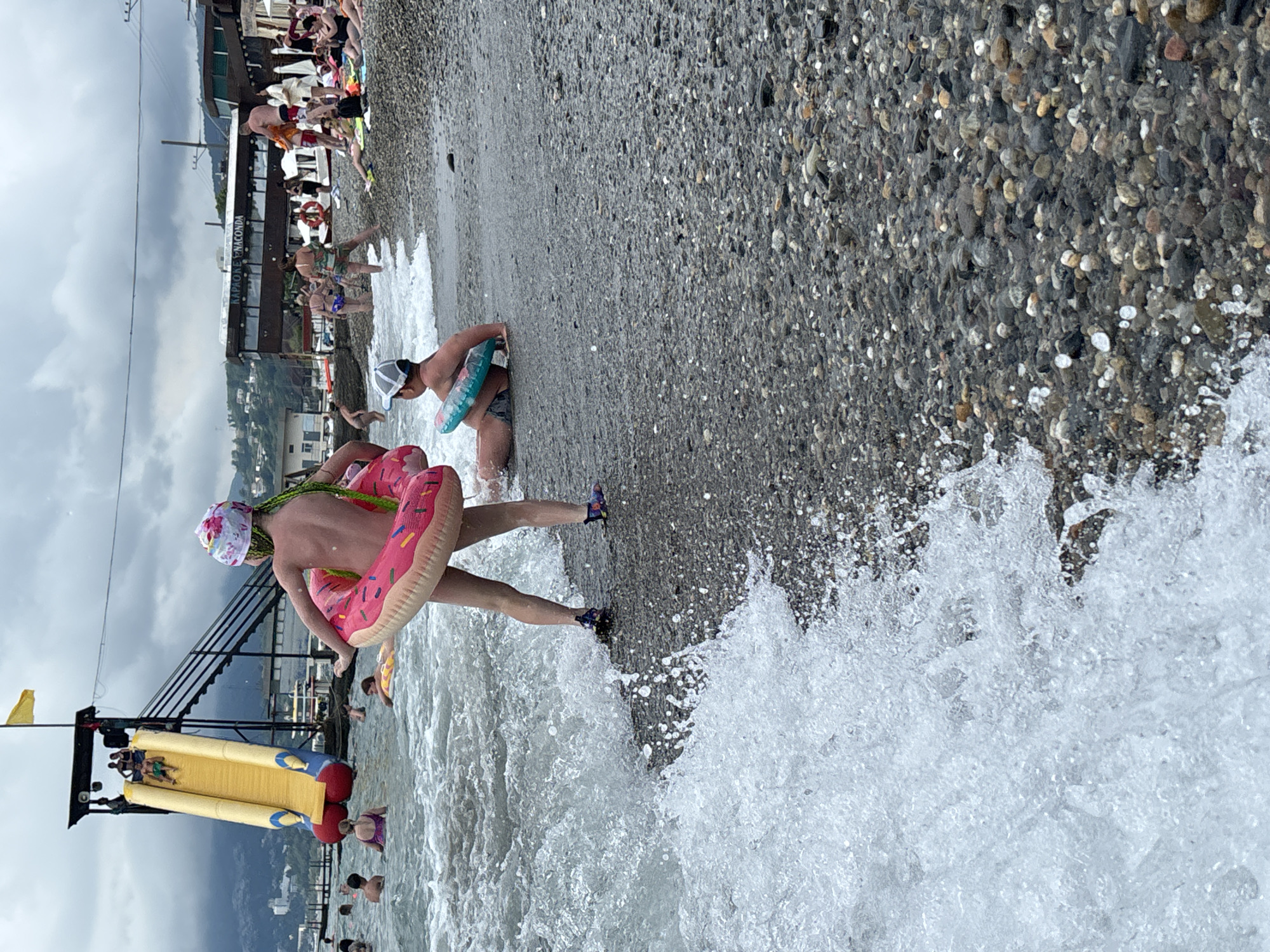 A group of people are enjoying a day at the beach on a cloudy day, likely in Sochi, Russia, given the presence of a large water slide and a building with signage indicating "Karaoke Anaconda". In the foreground, a child wearing a pink donut-shaped inflatable ring stands near the water's edge, facing away from the camera. To the right, another child sits on the pebbled shore with a teal inflatable ring. In the background, a yellow water slide is visible with several people on it or at the top. The sea is moderately wavy, with white foam crashing onto the shore. Other beachgoers are seen in the water and on the beach, some sunbathing, others swimming. The environment is a typical Black Sea resort beach with mountains in the distance.