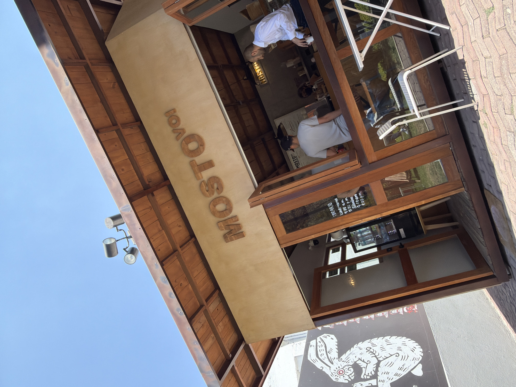 A slightly rotated, low-angle shot captures the exterior of the Mostovol restaurant located in San Andrés Cholula, Mexico, under a bright blue sky. The restaurant's name, "MOSTOVOL," is vertically printed in large wooden letters on the beige façade. The structure features a wooden roof overhang and wooden window and door frames.
Two individuals are visible inside. One man is wearing a white shirt, and another is wearing a light gray shirt and a baseball cap. Both are standing at an interior counter. Outside, folding white metal chairs are placed near the entrance.
The restaurant's operating hours are displayed on a sign near the window, and a white mural with a black rabbit graphic adorns the wall beneath the window. The exterior pavement is brick.