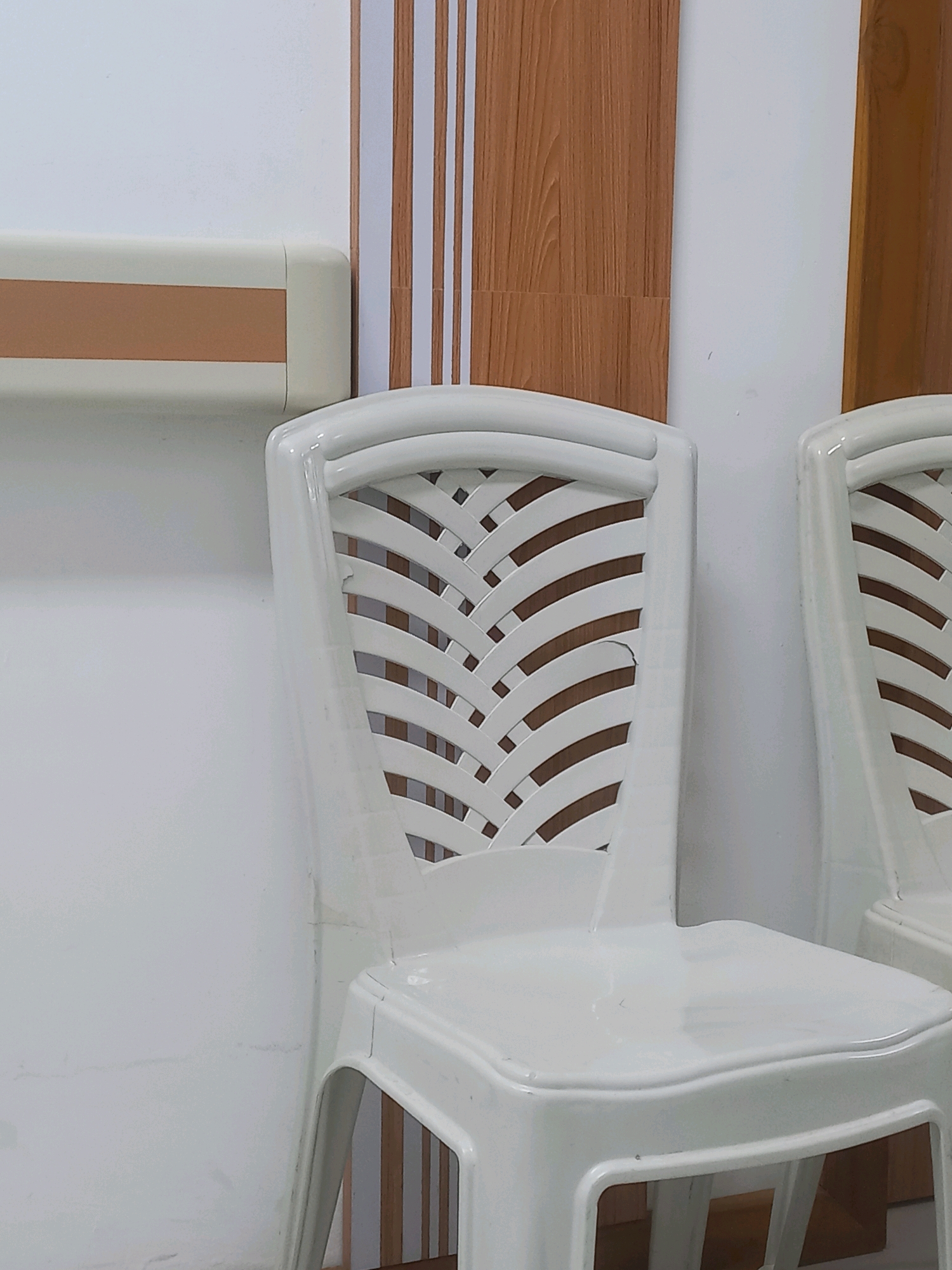 The image depicts an indoor scene, likely a waiting area or corridor, in Mergayu, Indonesia. In the foreground, two white plastic chairs are visible. The chair on the left is more prominently featured, showing a woven-patterned backrest and a smooth seat. The chair on the right is partially visible. The background consists of a white wall adorned with vertical wooden panels and a beige and brown handrail running horizontally along the wall on the left. The lighting suggests an indoor, possibly artificial, light source. There are no people or animals in the image.
