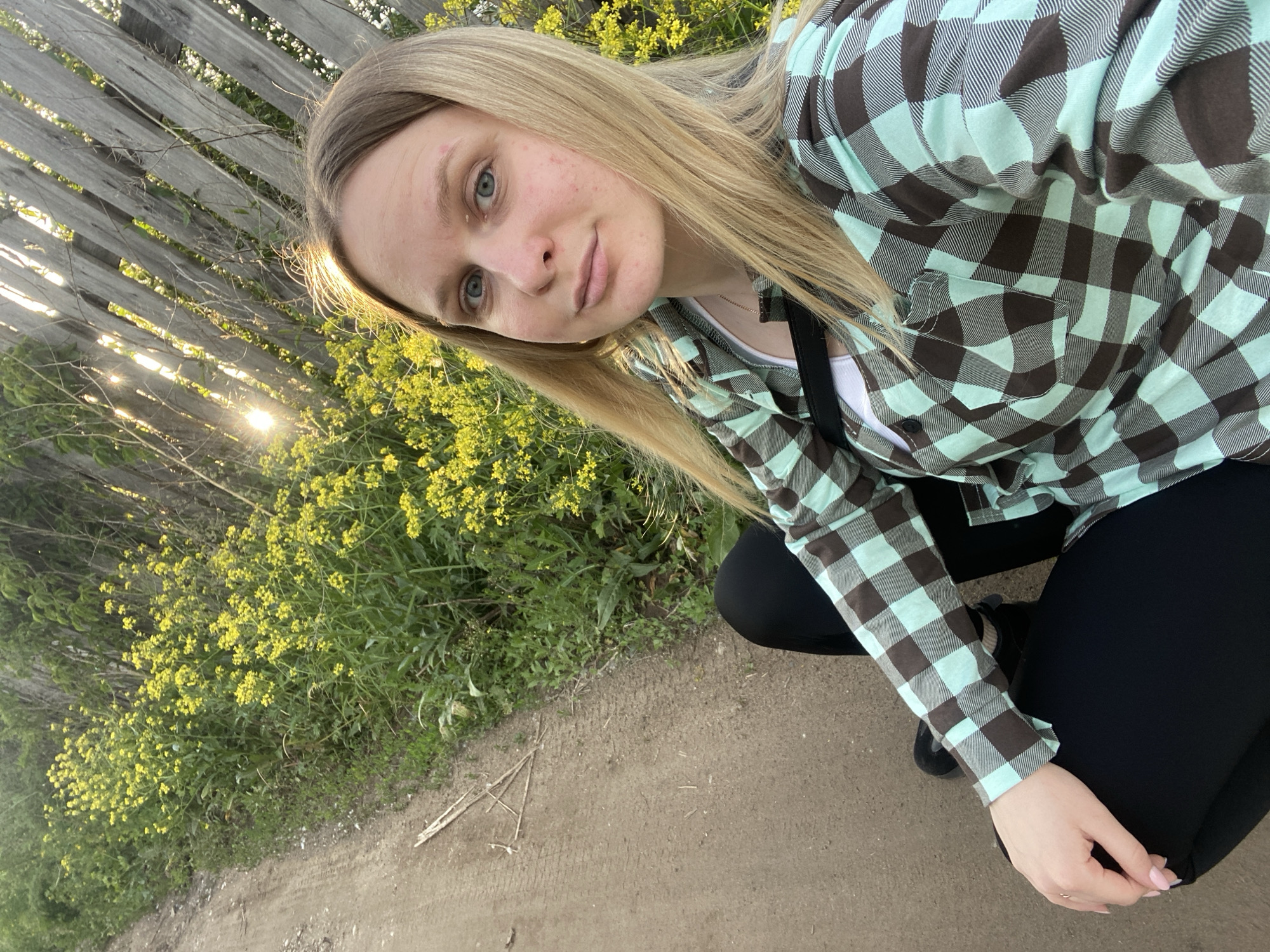 A young woman is depicted in an outdoor, angled photograph, likely taken as a selfie or close-up. She has long, straight blonde hair, blue eyes, and fair skin, and looks directly at the camera with a neutral, slightly contemplative expression. She is wearing a long-sleeved plaid shirt in light teal and brown over a white undershirt, and black leggings. Her left hand, with light pink fingernails, rests near her knee.
The woman is crouching or squatting on a dirt path. The background features a rustic wooden fence on the upper left, with abundant green foliage and clusters of small yellow flowers (resembling wild mustard or rapeseed) filling the middle and right sections. A bright sun is low in the sky, shining through the fence slats and creating lens flares, indicating late afternoon or early evening. The golden light illuminates the side of her face and backlights her hair. The scene evokes a rural or semi-rural outdoor setting, possibly in Chaikovsky, Russia, with clear, sunny weather. There is no visible text in the image.