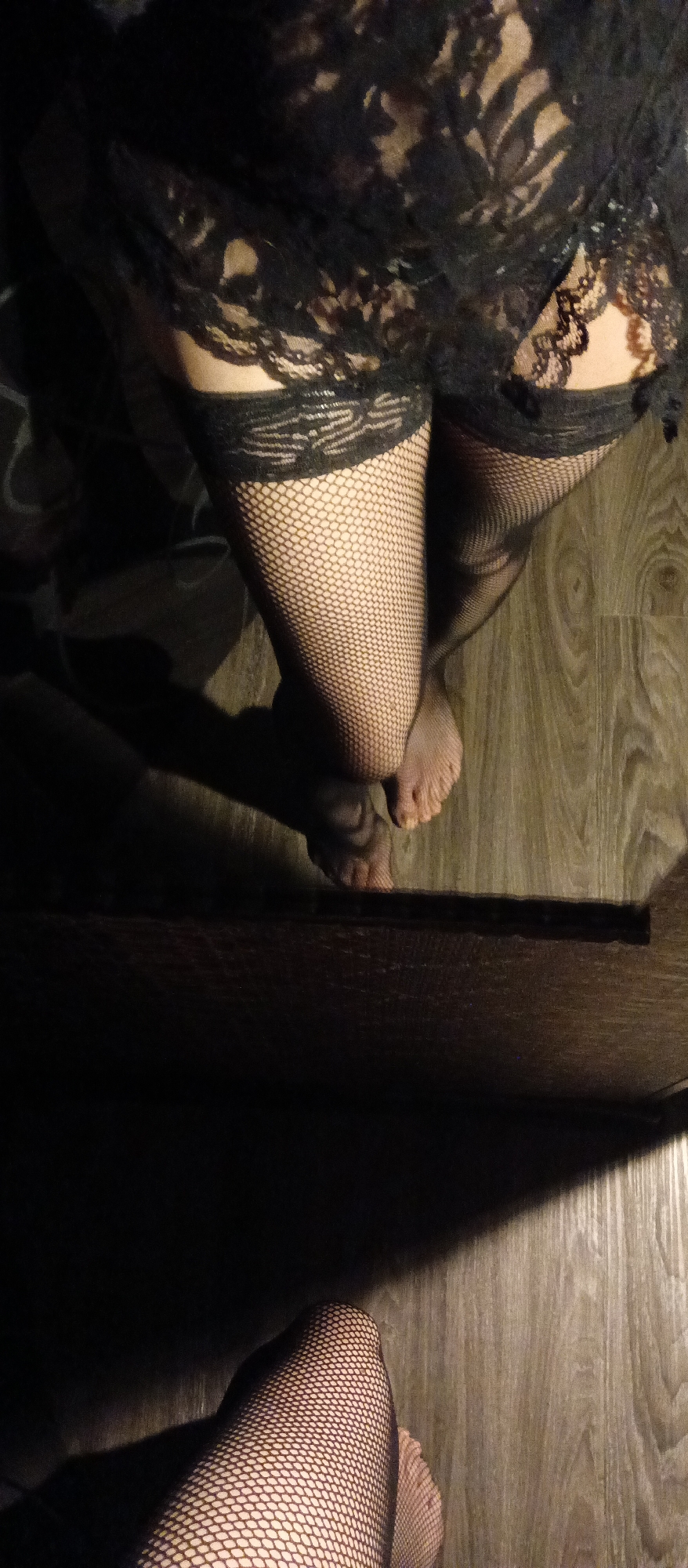 The image presents a view of a person wearing black fishnet stockings, with a lace garment partially visible. The person's feet are visible, bare, against a wood-grain floor. The lighting is subdued, creating strong shadows. The scene lacks specific contextual elements to determine a city or a forensic setting.