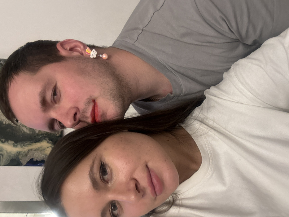 An indoor scene in Odintsovo, Russia, features two individuals posing for a photograph. In the foreground, a female with long, dark hair and light pink lipstick looks directly at the camera, wearing a white t-shirt. Behind her, a male with short, dark hair and light facial stubble is visible from the right side, wearing a grey t-shirt. He has red lipstick smudged on the left side of his mouth and cheek. A colorful earring, depicting a white cloud with a rainbow and a dangling pink/orange pearl-like bead, is present on his right ear. His expression is neutral. The background is blurred, showing a light-colored wall and a portion of an abstract art piece with dark and light swirls.