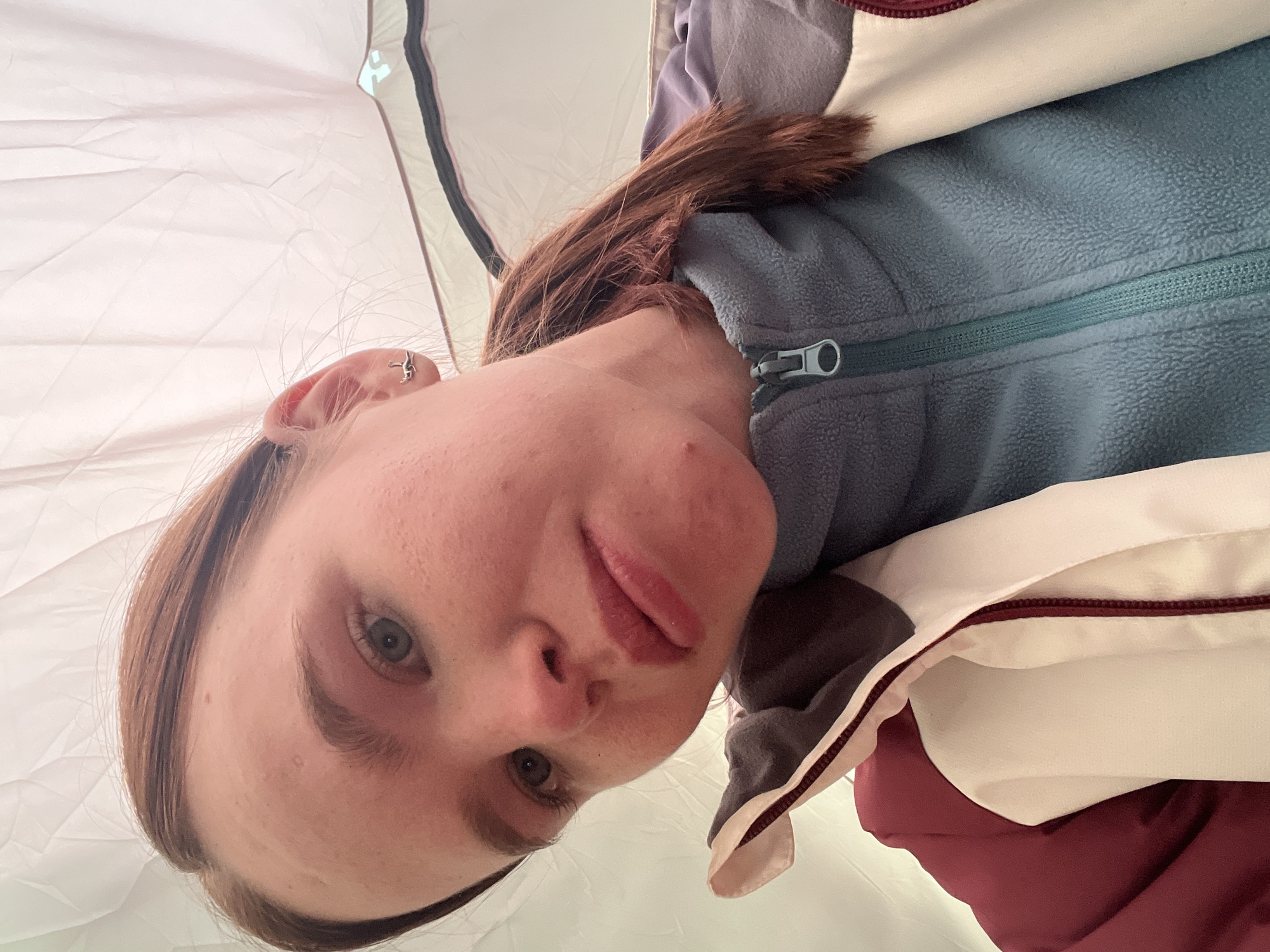 A close-up, rotated selfie captures a young person, likely female, with fair skin, light-colored eyes, and brown hair, looking directly at the camera. Their head is tilted, and their lips are slightly parted, revealing a hint of tongue. The expression is neutral to mildly curious.
The subject is wearing multiple layers of warm clothing, indicative of a cool environment. The most prominent inner layer is a blue-green fleece jacket with a visible zipper, worn over a purple-grey top. Over the fleece, there's a cream or off-white jacket with a maroon trim, which appears to be layered over another solid maroon garment. A small, silver-colored earring, shaped like a human figure in a dynamic pose, is visible on the helix of their left ear.
The background clearly depicts the interior of a tent, characterized by light-colored, wrinkled fabric walls. A dark, vertical strip on the right side suggests a tent pole or structural seam. The lighting is soft and diffused, consistent with being inside a tent or during an overcast day. The overall scene conveys a sense of outdoor activity or camping, potentially in a cooler climate in Lesnoye, Russia, given the clothing choices and setting. No specific time of day or weather conditions are discernible, nor is any visible text present in the image.