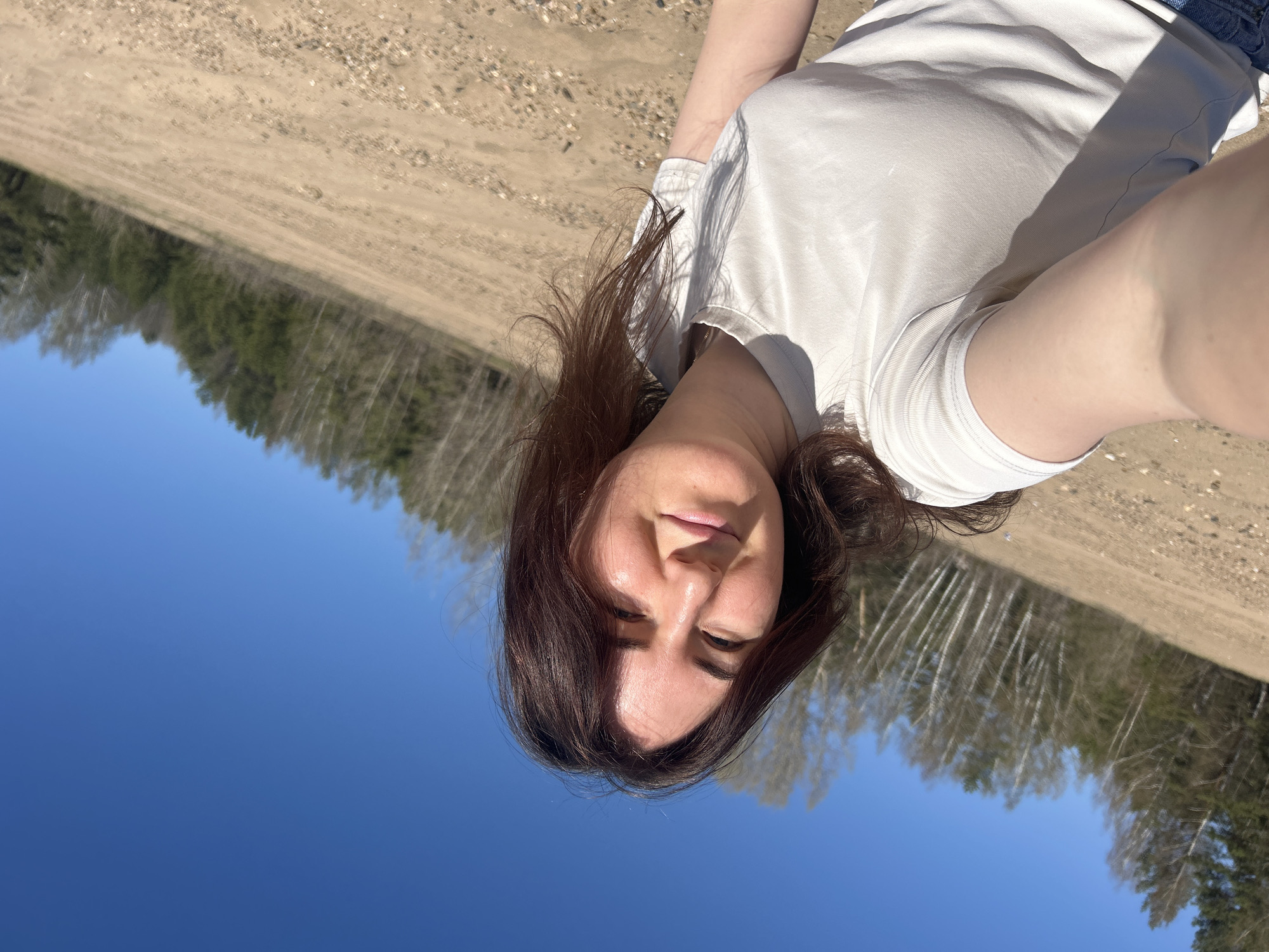 A woman is taking a selfie outside in what appears to be a natural environment, possibly on the outskirts of Noviy, Russia. The background includes a clear blue sky and a forested area. The ground appears to be sandy or gravelly. The woman has brown hair and is wearing a white t-shirt. She is facing the camera with a slight smile.