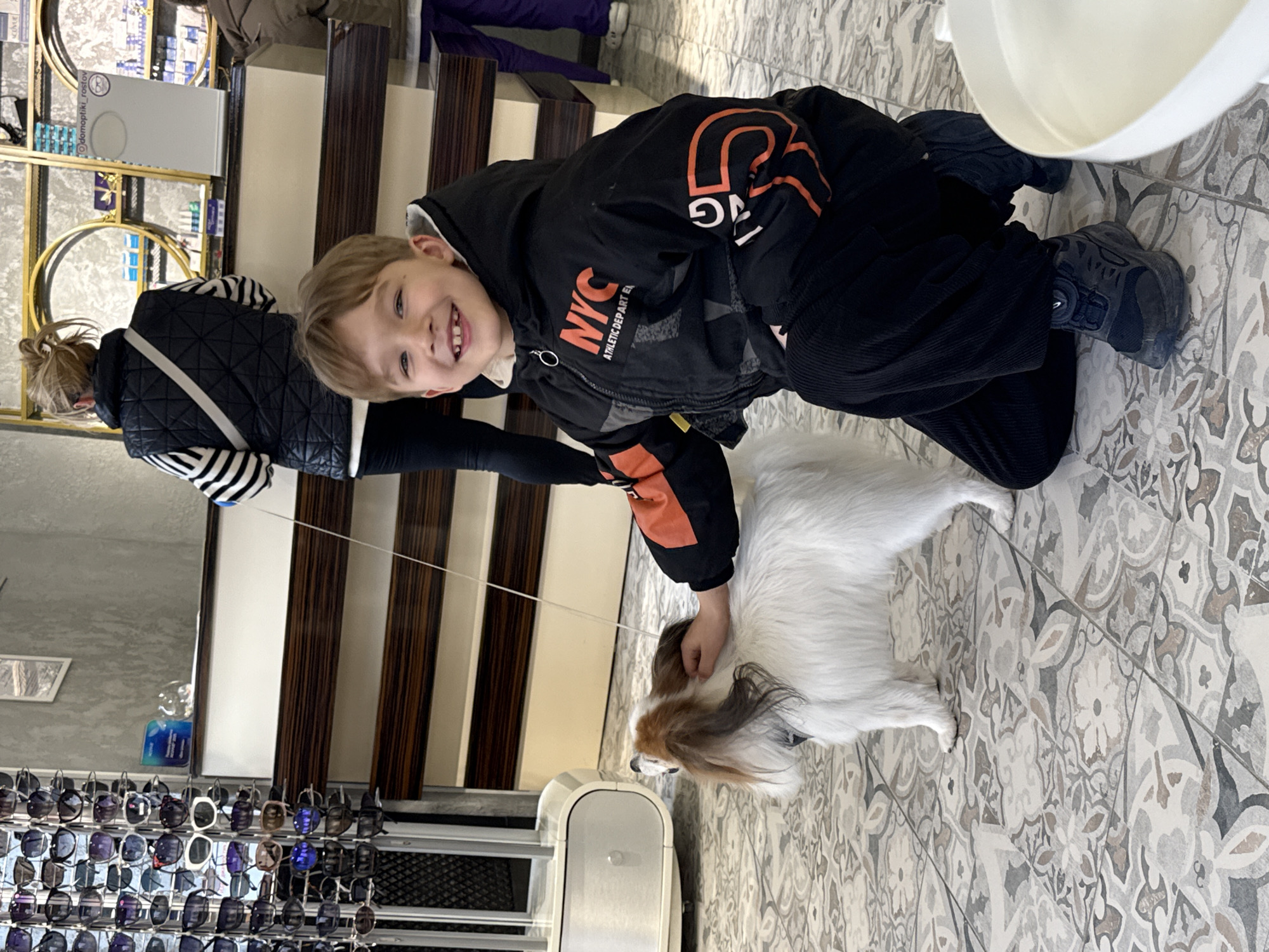A young boy in a black jacket is kneeling on a patterned tile floor, petting a white and tan Papillon dog. The boy smiles at the camera. Behind him, a woman in a black and white quilted vest and a white crossbody bag is walking, holding a leash. A set of wooden and white stairs rises behind the woman. To the left, a display of sunglasses is visible. In the background, shelves with items are visible. The location is likely a retail store in the city of Ovoshchnoy, Russia, suggested by the text visible on the shelf, "domoptiki, rostov".