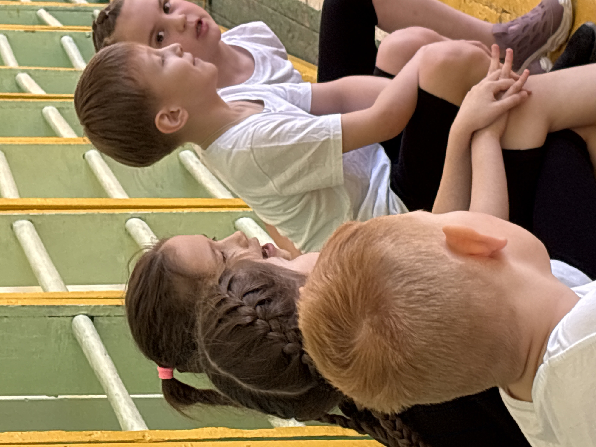 Four children are sitting on a yellow and green staircase. Three are visible from the waist up, and one from the neck up. All the children have pale skin. The child at the top is looking up and to the left, wearing a white t-shirt and black leggings. The child next to them is looking up and to the left, wearing a white t-shirt. The child next to them has their hair braided and is wearing a white t-shirt. The child at the bottom has red hair. The image shows people in Izhevsk, Russia.