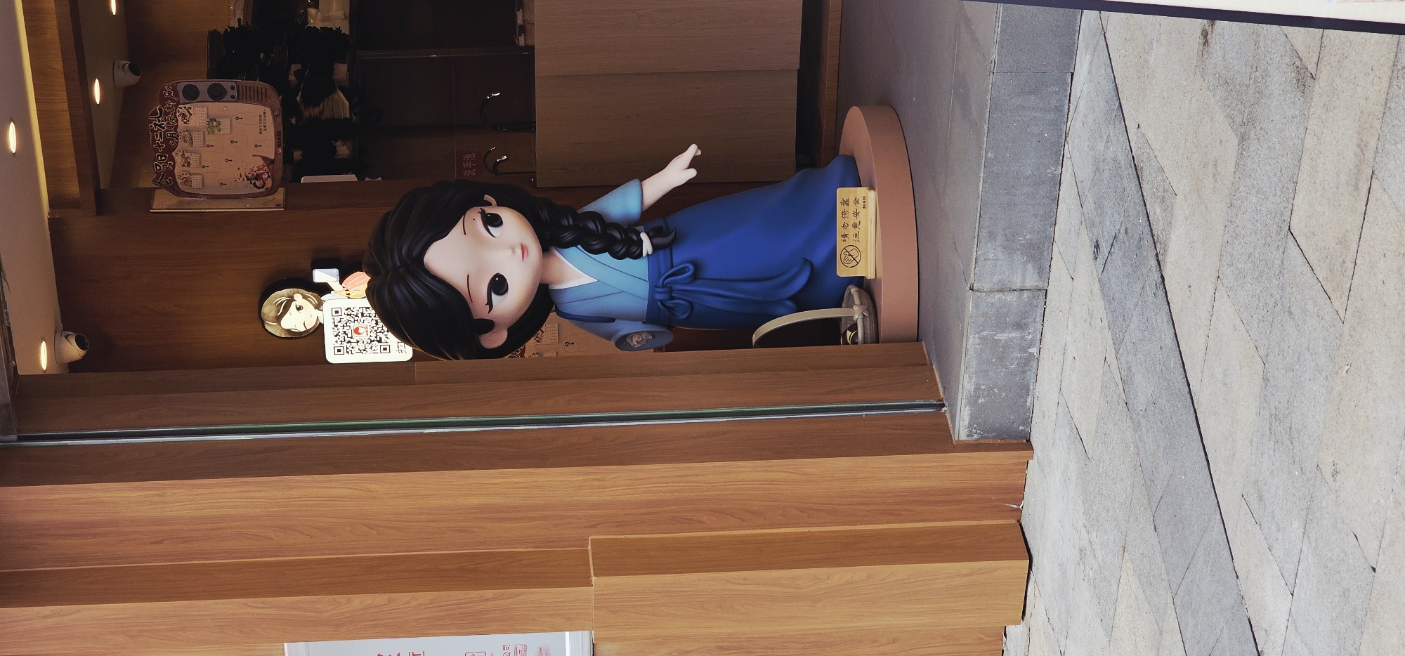 The image shows a storefront in what appears to be the Chongqing High-tech Zone in China, judging by the Chinese characters on signs. A cartoonish statue of a girl with black hair in a blue traditional dress is positioned on a round stand just inside the entrance. A sign on the stand contains Chinese text. The statue is in front of the shop's wooden exterior and a glass door. Inside the store, a display board with food items and prices, along with a QR code, are visible. Outside, on the right side of the frame, is a paved walkway with large, irregularly shaped stones.
