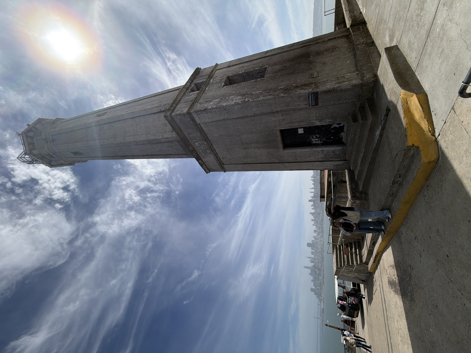 In a wide-angle, low-angle shot, a concrete lighthouse stands on Alcatraz Island in San Francisco Bay, United States of America. The lighthouse, with its octagonal shape and weathered surface, is positioned on the right side of the frame, angled slightly towards the viewer. The top of the lighthouse is capped with a metal structure. The sun shines brightly in the upper right portion of the sky, casting a lens flare and visible rainbow effect. Wispy clouds streak across a clear blue sky. In the background, the cityscape of San Francisco is visible across the water, with numerous skyscrapers and buildings forming a dense urban skyline. The foreground consists of a concrete walkway and stairs leading up to the lighthouse. Two women are standing on the stairs in the middle ground, looking towards the camera. To the left of the lighthouse, a trolley or bus is parked, with several people standing near it and a few more seated inside. The overall scene depicts a tourist location on a sunny day with the iconic San Francisco skyline in the distance.