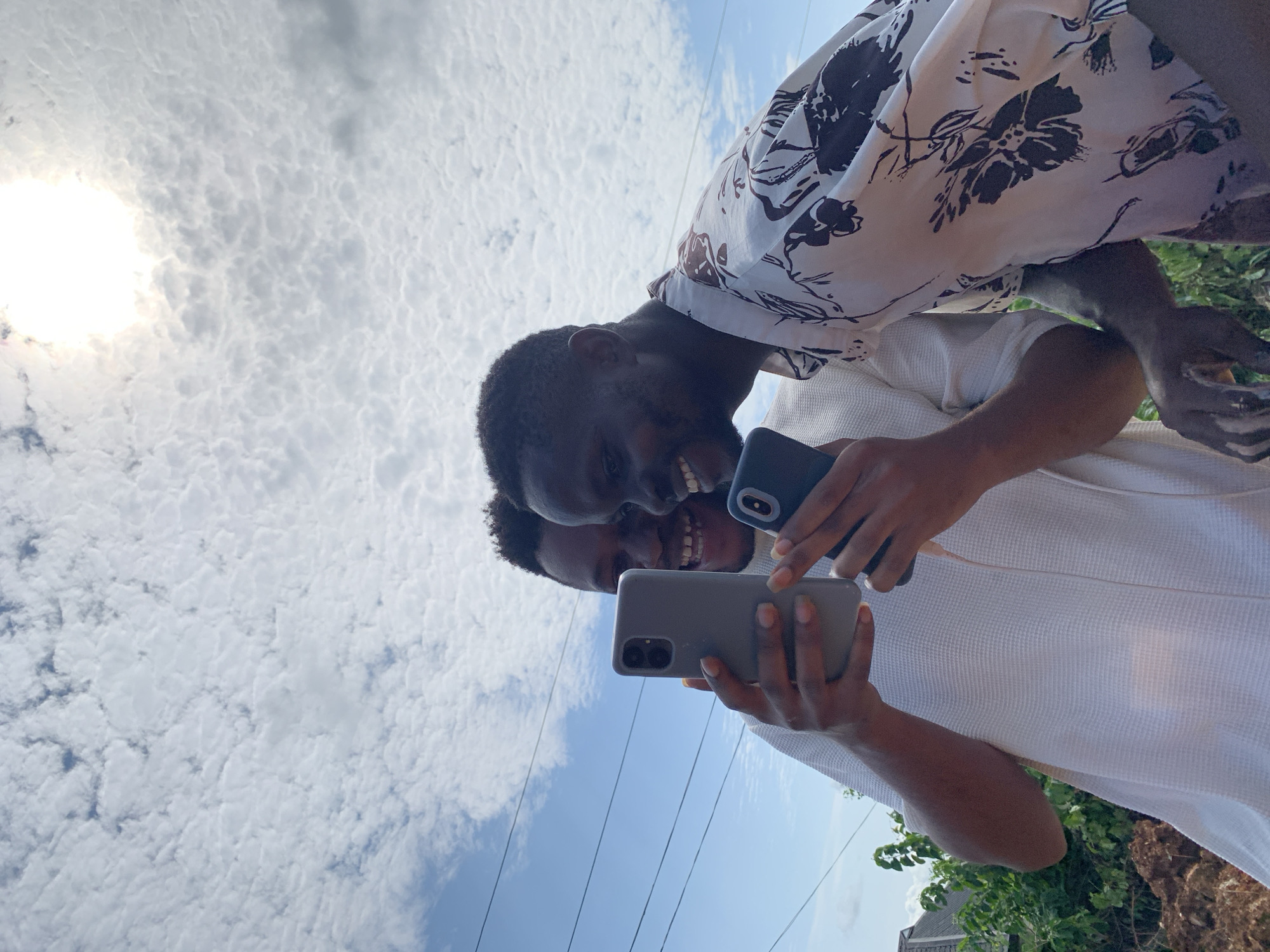 Two young men are taking a selfie outdoors under a bright sky. The man in the foreground is wearing a white waffle-knit shirt and is holding a grey smartphone in his right hand, showing its dual rear cameras. He is smiling broadly, looking towards the phone as if posing for the picture. Behind him and slightly to the left, another man, wearing a white shirt with a dark floral pattern, is also visible. He is leaning in, also smiling and looking at the phone. He is holding a dark blue smartphone in his left hand.
The sun is visible in the upper left corner of the image, suggesting it is daytime. The sky is filled with scattered, textured white clouds against a bright blue backdrop. In the lower portion of the image, electrical wires are visible stretching horizontally, indicating an urban or semi-urban environment. Green foliage and some brown earth are visible on the right side, hinting at an outdoor setting with vegetation. The overall mood appears joyful and casual, with both individuals engaged in capturing a moment together. The image is oriented vertically, with the subjects appearing upside down relative to a typical ground-level perspective.