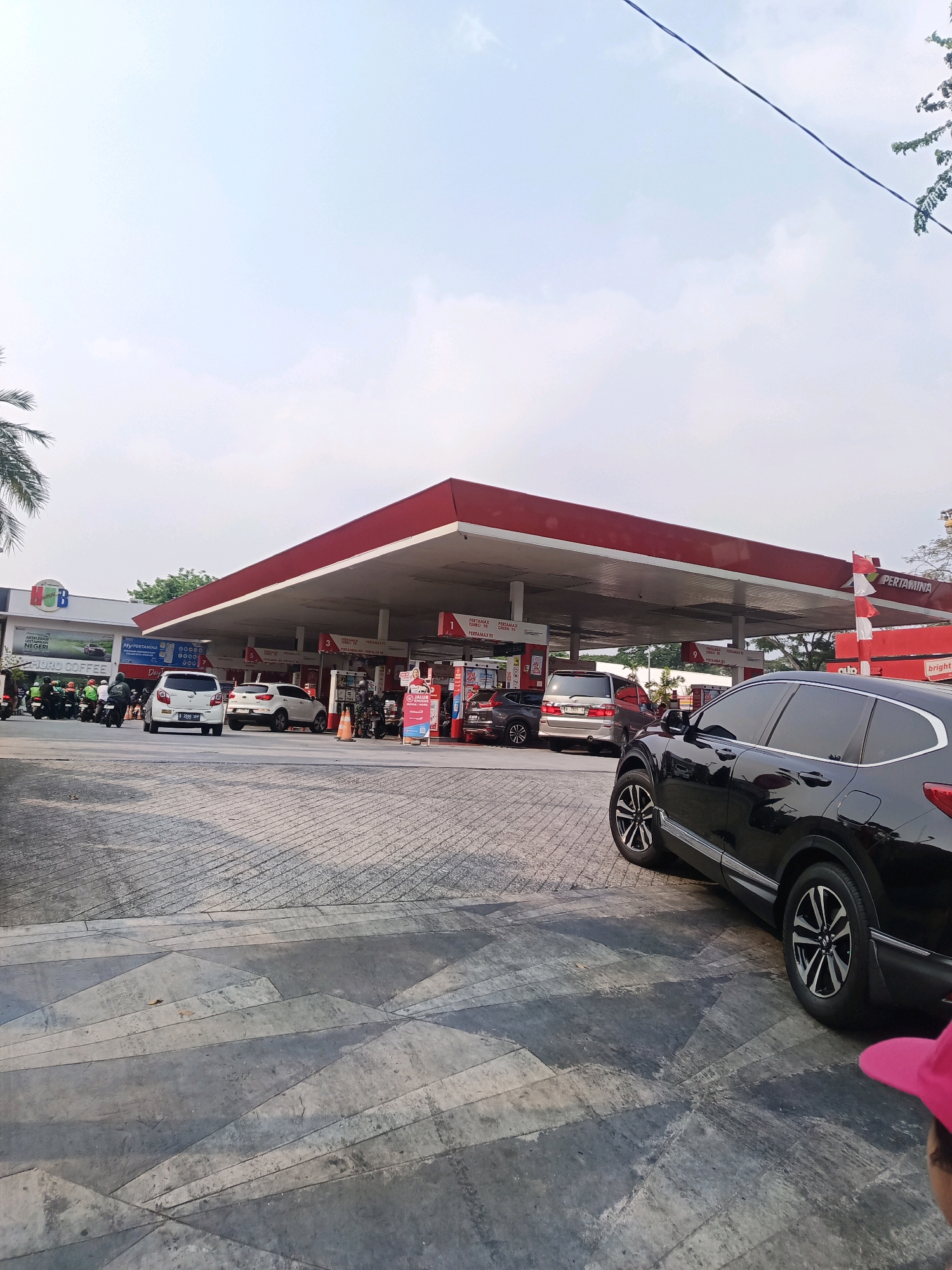 The image shows a busy Pertamina gas station in South Jakarta, Indonesia, on a clear day. Several vehicles, including cars and motorcycles, are lined up at the pumps. A black SUV is parked in the foreground, obscuring part of the scene. The gas station has a red roof supported by white pillars, and signs indicate various fuel types. A convenience store branded "Indomaret" is visible to the left of the gas station, with a sign that reads "MORO COFFEE". The ground is paved with a patterned concrete surface. The sky is blue with scattered clouds.