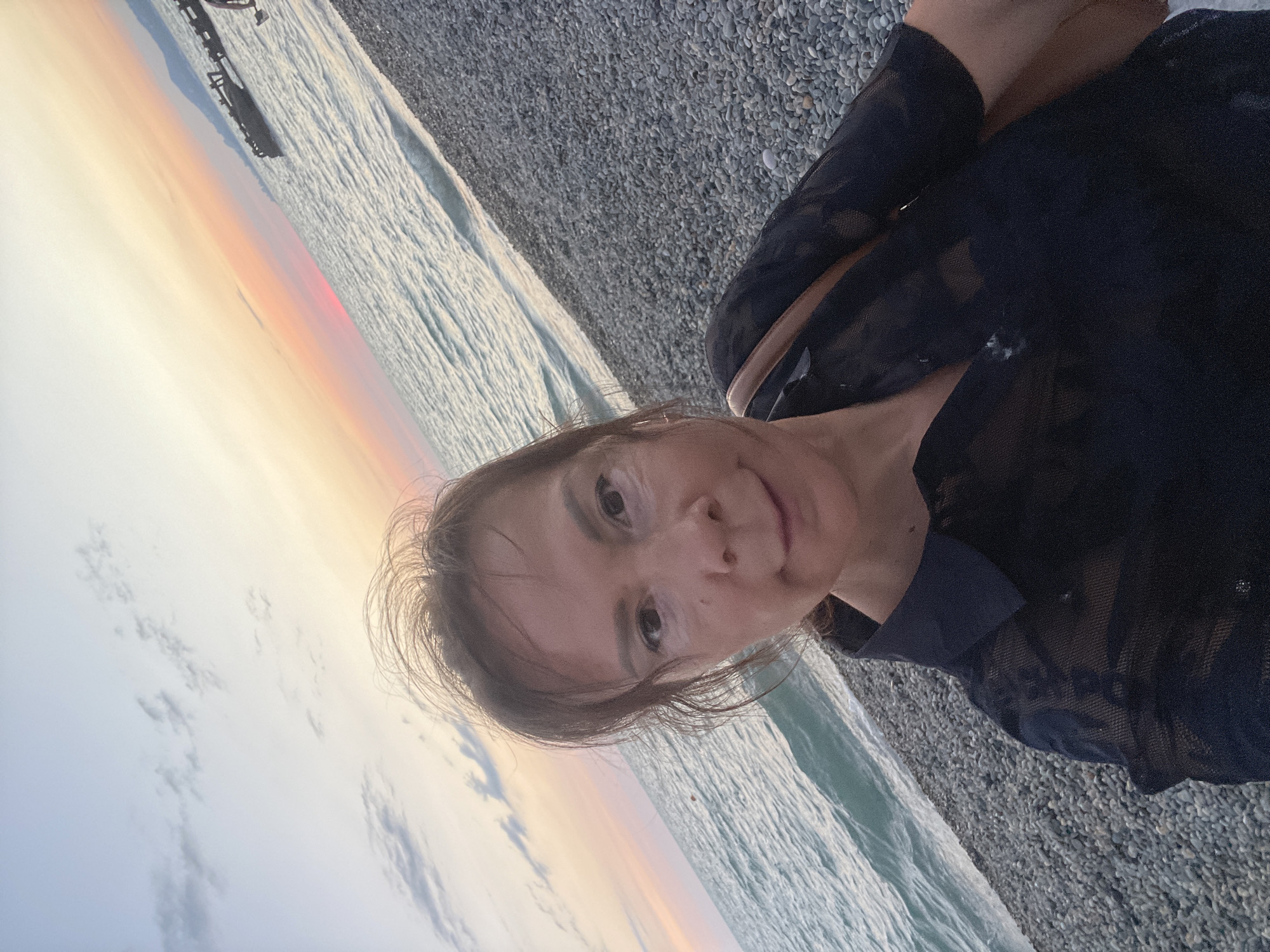 A medium-shot selfie shows a woman with medium brown hair and light brown eyes taking a picture of herself on a pebbled beach at sunset. The woman is wearing a black lace shirt and a tan purse is slung over her left shoulder. The ocean is to her left with gentle waves. A pier is visible in the background on the right side of the image. The sky is a mixture of pale blue and orange, indicating that the sun is setting. The overall scene is peaceful and serene. The location is the city of Sochi, Russia.