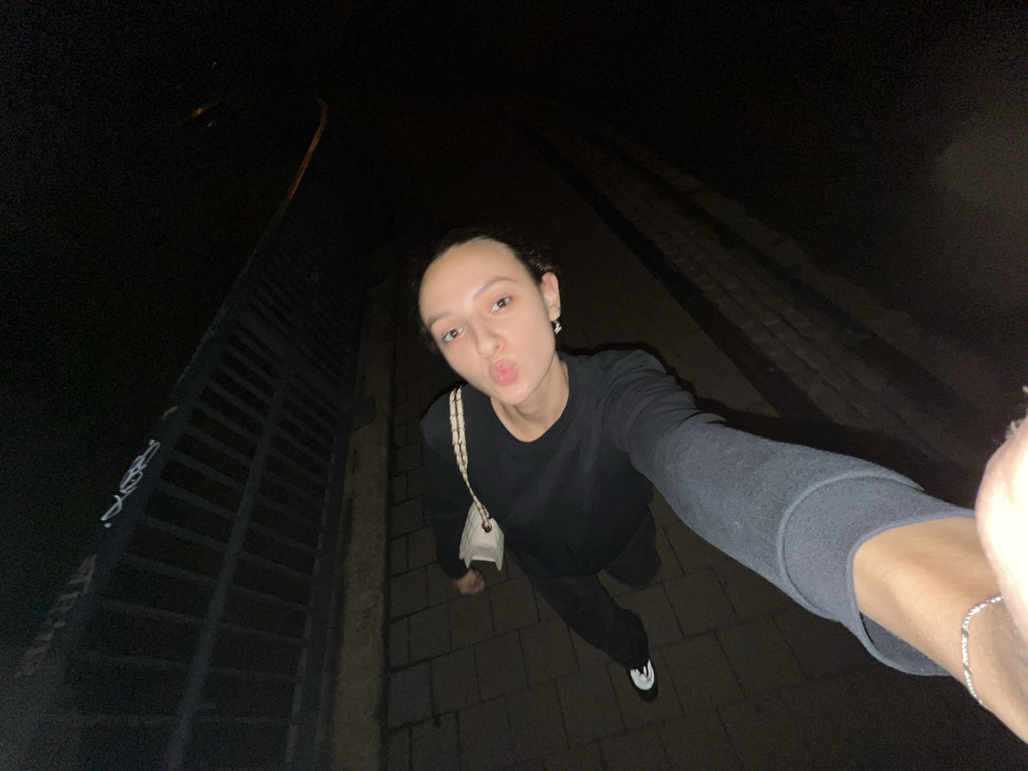 A young woman is captured in a wide-angle, low-light selfie taken outdoors at night in Saarbrücken, Germany. She stands centrally, looking upwards and directly at the camera with a playful "duck face" or puckered lips expression. Her dark hair is pulled back, revealing her face, and she wears a simple black long-sleeved top and dark trousers. A light-colored, possibly white or cream, quilted shoulder bag with a gold-toned chain strap is slung over her left shoulder. She accessorizes with small earrings and a silver chain bracelet on her right wrist, which is extended towards the camera, indicating she is holding the phone for the selfie. Her right foot is visible, wearing a black sneaker with a distinct white toe cap and sole.
The background is predominantly dark, characteristic of a night scene. The ground beneath her is paved with dark, rectangular blocks, resembling a sidewalk or pathway. To the left of the frame, a dark, slatted metal railing or fence extends diagonally into the background. On this railing, light-colored graffiti is visible, including what appears to be stylized Arabic script, with discernible words like "توب" (top) and "خواب" (dream). Faint, blurry streetlights or distant lights can be discerned in the upper right portion of the background, adding to the urban night atmosphere. The image has a slightly grainy texture due to the low light conditions. The overall mood is casual and spontaneous.