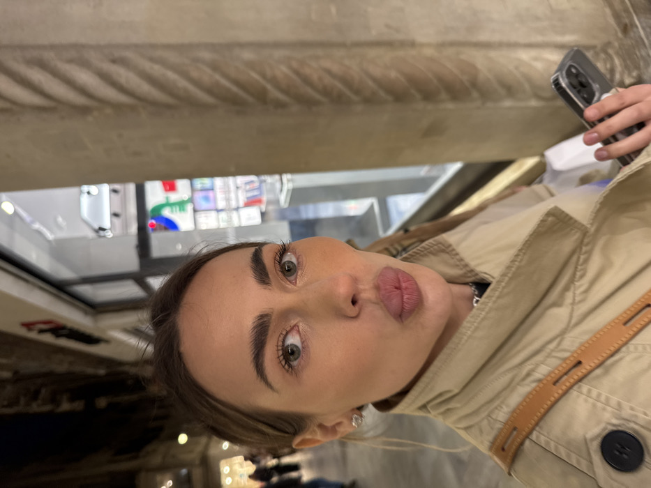 A woman is depicted in a close-up selfie in a nocturnal urban environment, identified as Venice, Italy. She has light-colored eyes, dark eyebrows, and her lips are puckered. Her light-brown hair is pulled back, and she wears a beige trench coat with a visible dark button and a brown leather strap from a bag across her shoulder. A silver stud earring is visible in her left ear. Her left hand holds a dark smartphone in a clear case, with a triple-lens camera array visible on its back. To her right, a prominent, light-colored stone column with a distinctive twisted, fluted design stands beside a retail storefront with a large glass display window. The store exhibits illuminated products, likely electronics, and features a brightly colored logo with red and green components. The background to the left shows blurred lights and indistinct figures under what appears to be an illuminated pedestrian arcade. The architectural characteristics, particularly the twisted column and arcaded setting, are consistent with the Procuratie Vecchie or Nuove in Piazza San Marco, Venice.