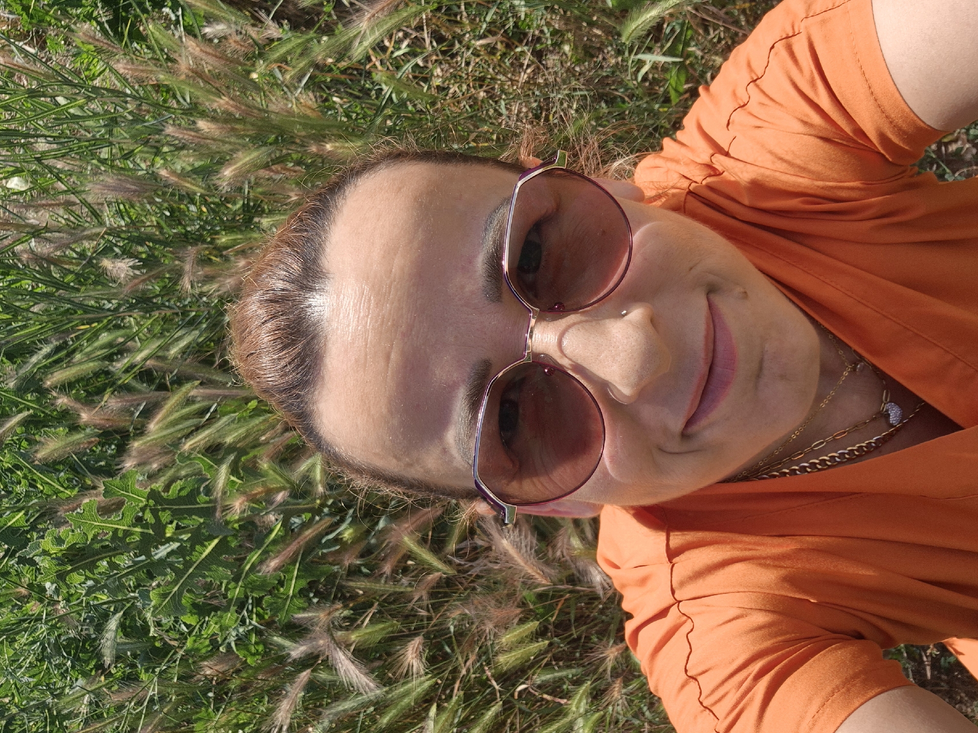 The media file is a close-up, overhead shot of a woman lying on the grass, possibly in a park or field. The woman is wearing an orange t-shirt and sunglasses. She has her eyes closed and a slight smile on her face, suggesting she is relaxed and enjoying the moment. The sun appears to be shining, casting shadows and highlighting the texture of the grass and her skin. The woman has a nose piercing and is wearing a few necklaces, one of which has a small, sparkly pendant. The image is taken from a high angle, looking down at the woman. The grass around her is green and lush, with some dried seed heads mixed in, indicating it might be late spring or summer. The lighting suggests it is daytime, likely afternoon, with the sun creating a warm glow.