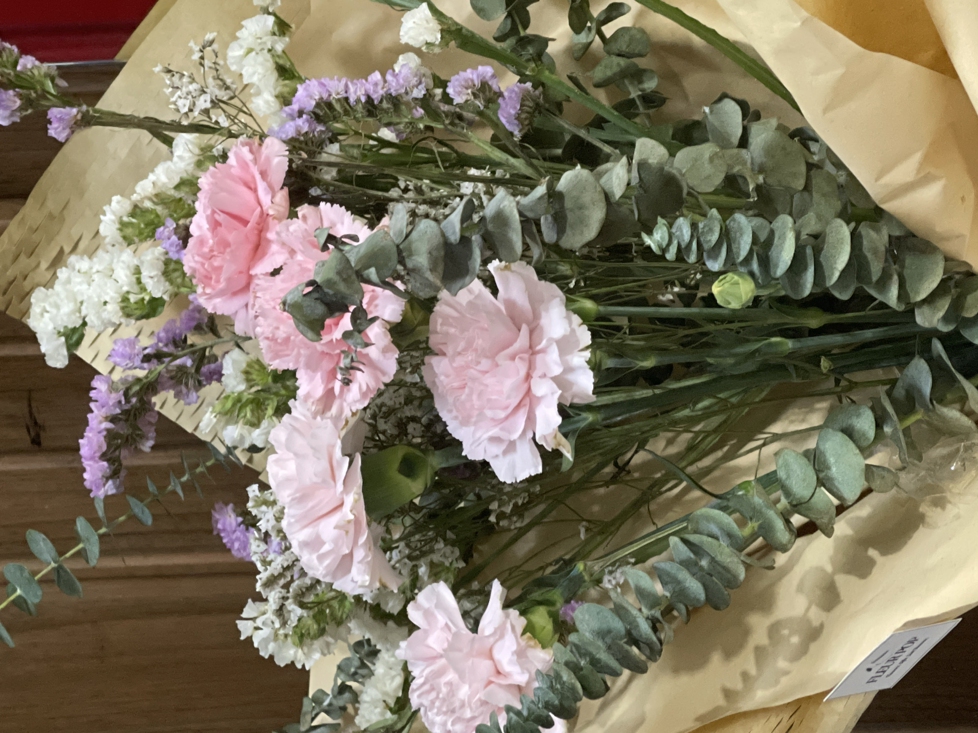 A bouquet of light pink carnations, white and lavender statice flowers, and eucalyptus leaves are arranged in brown paper. The scene is indoors, with a wooden surface visible in the background. There is a small white tag with text on it located at the bottom right of the paper. The location is in Davao City, Philippines.
