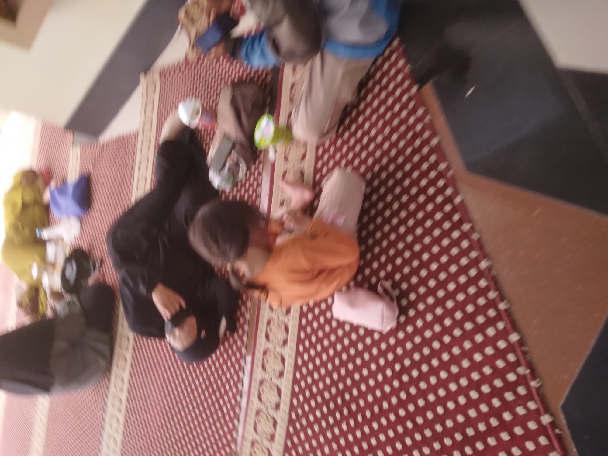 In Desa Pamijen, Kecamatan Sokaraja, Indonesia, a group of people are seen resting on a red carpet with a white geometric pattern. The scene appears to be indoors, possibly a mosque or a community hall, given the attire and the presence of prayer rugs. A woman in a black hijab and abaya is lying down, while another individual, also in dark clothing and a hijab, is seated nearby. A young girl in an orange shirt is sitting on the floor, holding a small pink bag. Another person, partially visible, is dressed in a blue jacket and beige pants. Several objects are scattered around, including plastic cups, a water bottle, and what looks like a rolled-up prayer mat. The lighting is dim, casting soft shadows across the room. The overall impression is one of a tranquil gathering, with individuals relaxing or taking a break.