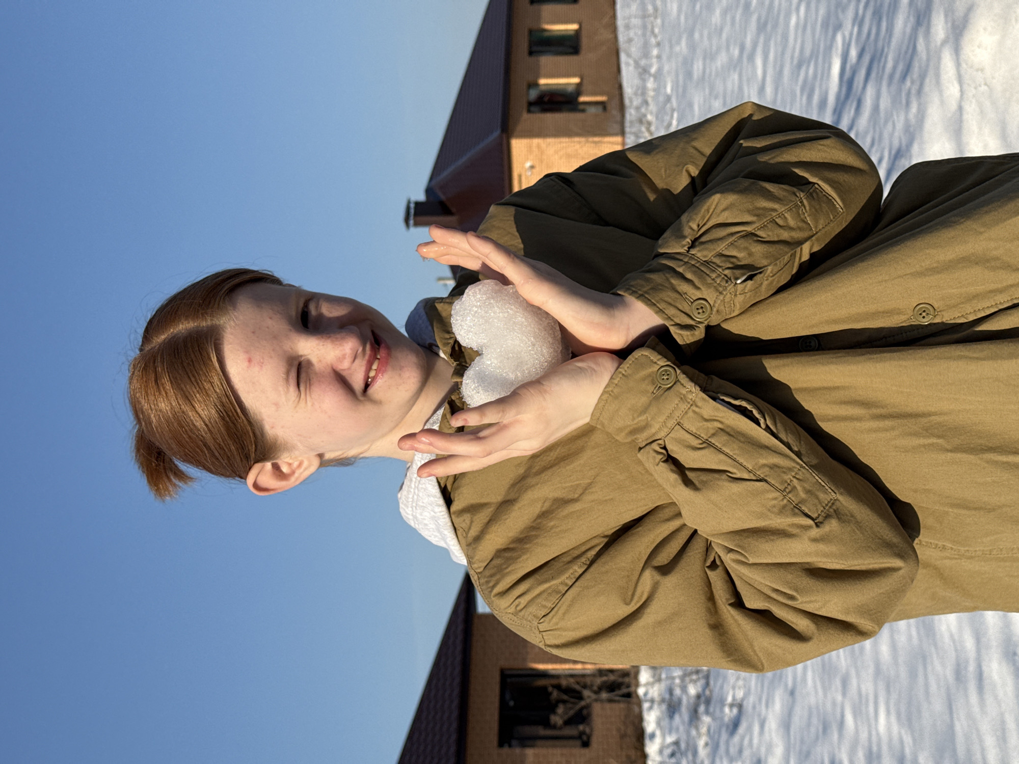 A young person with reddish-brown hair, styled in a ponytail, stands in a snow-covered field in Chaikovsky, Russia. They are wearing a light brown collared jacket over a grey hooded sweatshirt. The individual smiles and winks with their left eye while holding a heart-shaped snow sculpture in cupped hands. The background features a clear blue sky and two single-story brick houses with dark brown gable roofs. The ground is uniformly covered in snow, with shadows cast by visible and unseen objects under bright sunlight.