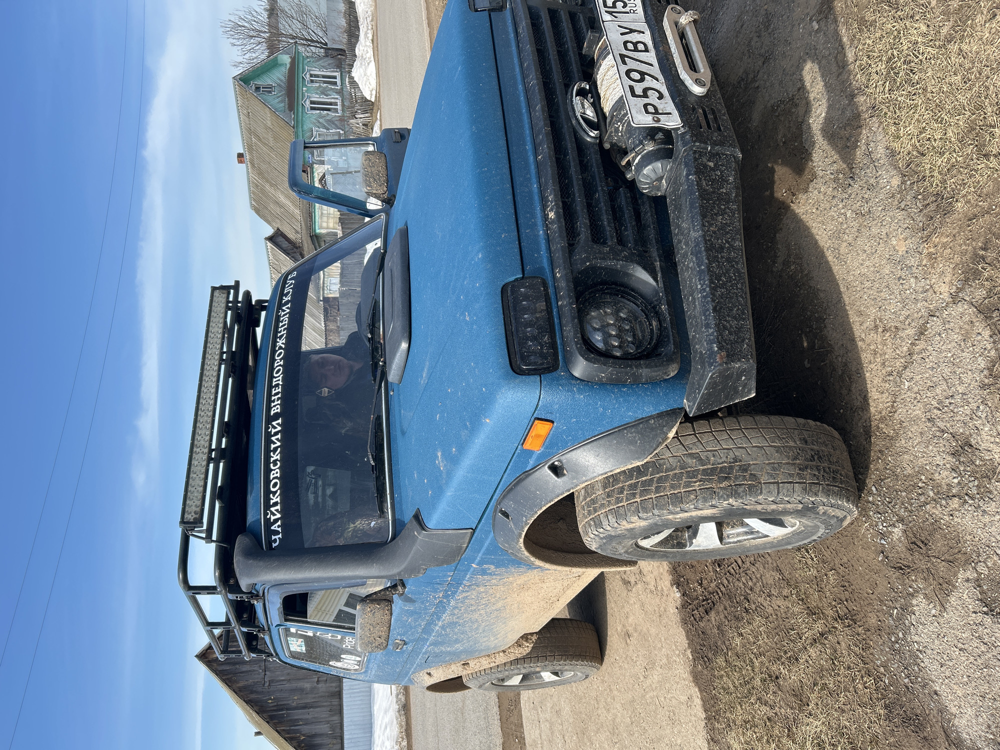 The image depicts a blue off-road vehicle, likely a Lada Niva, parked on a dirt and gravel surface. The vehicle is covered in mud, suggesting recent off-road use. The front of the vehicle is visible, showing a black grille, LED headlights, a winch, and a license plate with the registration number "P597BY 15 RUS". A snorkel is mounted on the driver's side. There is an aftermarket roof rack with a light bar. The phrase "ЧАЙКОВСКИЙ ВНЕДОРОЖНЫЙ КЛУБ" (Chaikovsky Off-Road Club) is written on the windshield header.
In the background, there are wooden buildings typical of rural Russian architecture, including a two-story house with a gable roof and painted windows. The presence of the phrase “Чайковский Внедорожный Клуб” suggests the location is near the town of Chaikovsky, which is within the larger geographic location of Уральское, Russia. The sky is blue with some clouds, indicating daytime. The environment suggests a rural setting, possibly on the outskirts of a town or village.