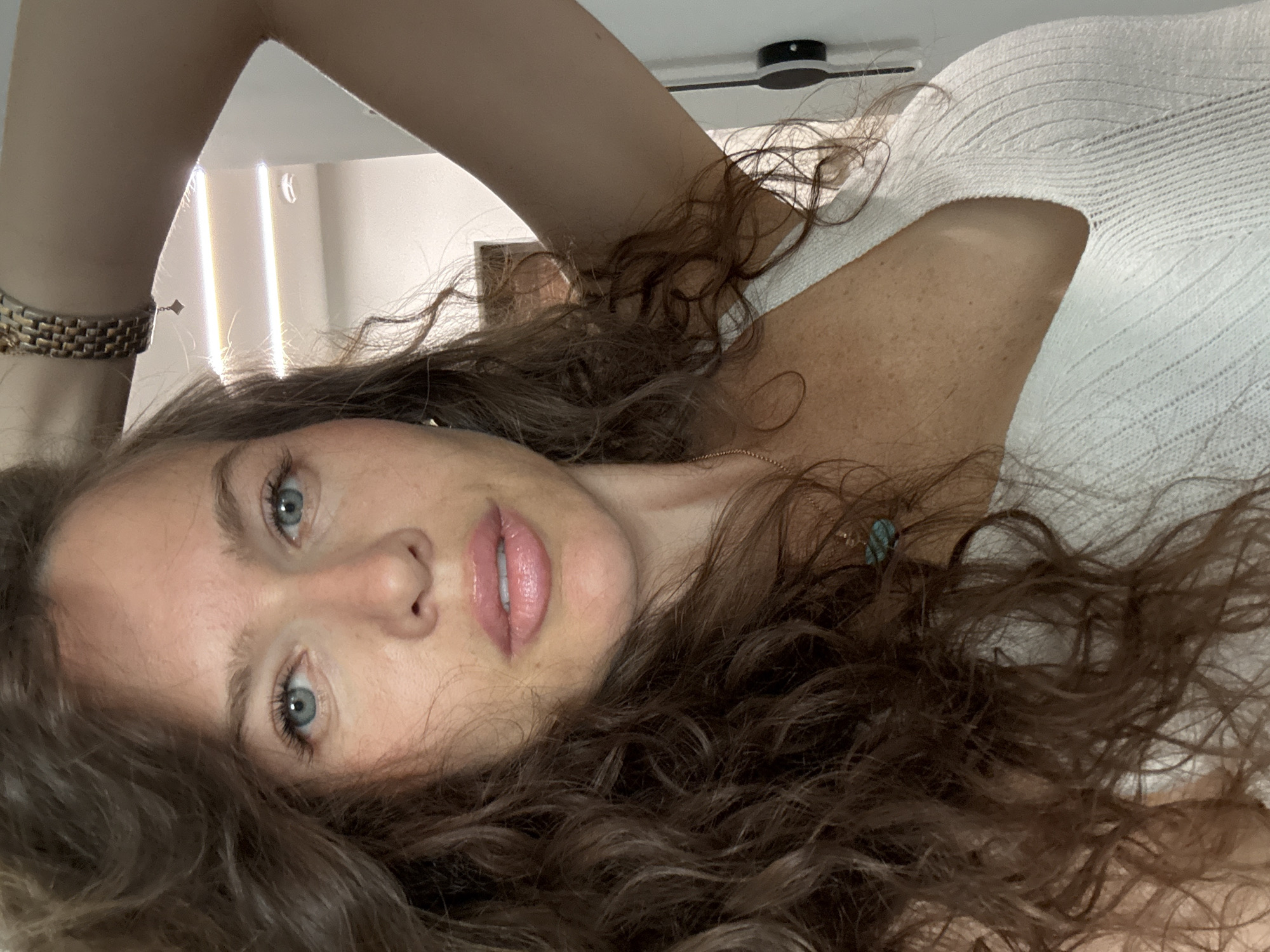 A woman with long, curly brown hair and blue eyes is captured in a close-up shot. She is wearing a white, ribbed tank top. Her right arm is raised, with her hand resting on her head. A gold-toned watch with a textured band is visible on her wrist. She is also wearing a delicate gold necklace with a small, turquoise-colored pendant. The background is slightly blurred, showing a white wall with overhead lighting fixtures and what appears to be a doorway or alcove. There are no discernible features that would indicate the location in the city of Khimki, Russia.