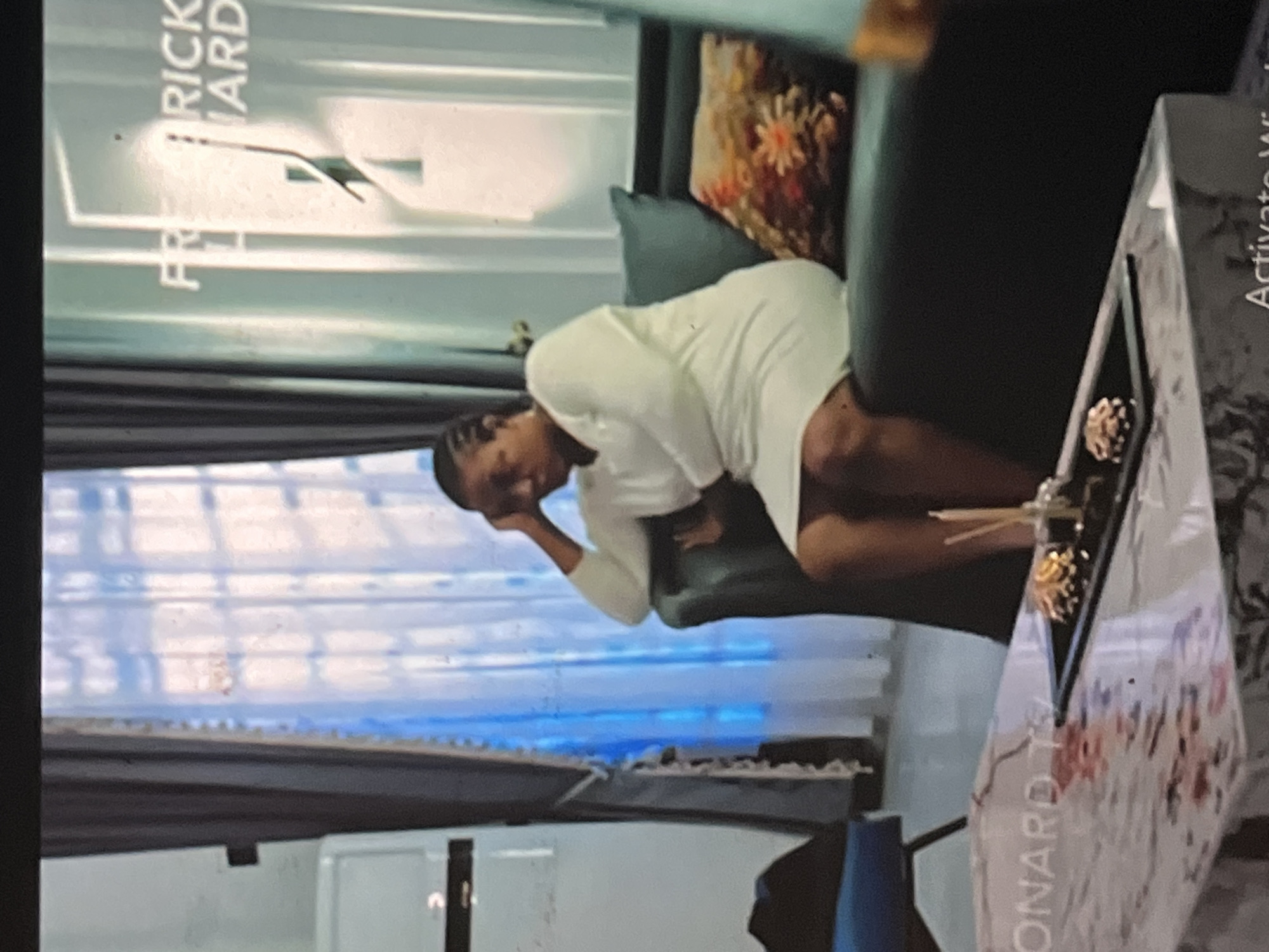 A woman in a white dress sits on a green couch in a room. Behind her is a window with a blue curtain. In front of her is a coffee table with gold-colored decorative elements. The TV shows the name "FREDRICK WARD" and "LEONARD TV," indicating the program is likely filmed or based in the city of Abuja, Nigeria.