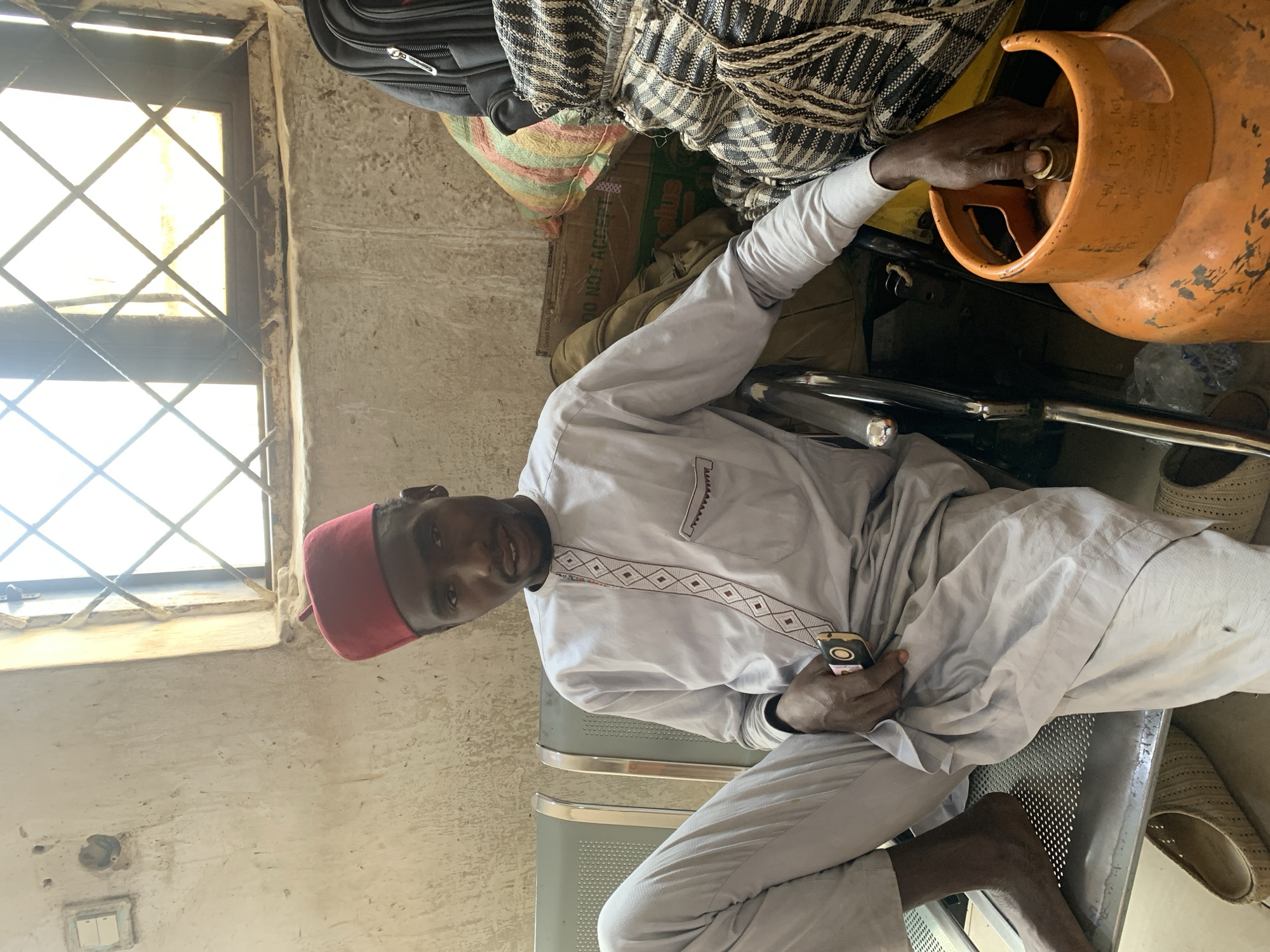 A man sits indoors, likely in the city of Jos, Nigeria. He wears a light-colored traditional tunic with embroidered detail on the chest, gray pants, and a red fez. He holds a cell phone in one hand and reaches toward an orange propane tank with the other. The tank rests on a black metal frame. Behind him are several bags and suitcases stacked on top of a cardboard box labeled "DO NOT ACCEPT IF SEAL IS BROKEN." He is sitting on a gray metal chair. A window with a diamond-shaped grill is visible in the background. The walls are painted in a weathered off-white color. An electrical outlet and light switch can be seen on the wall to the left of the window.