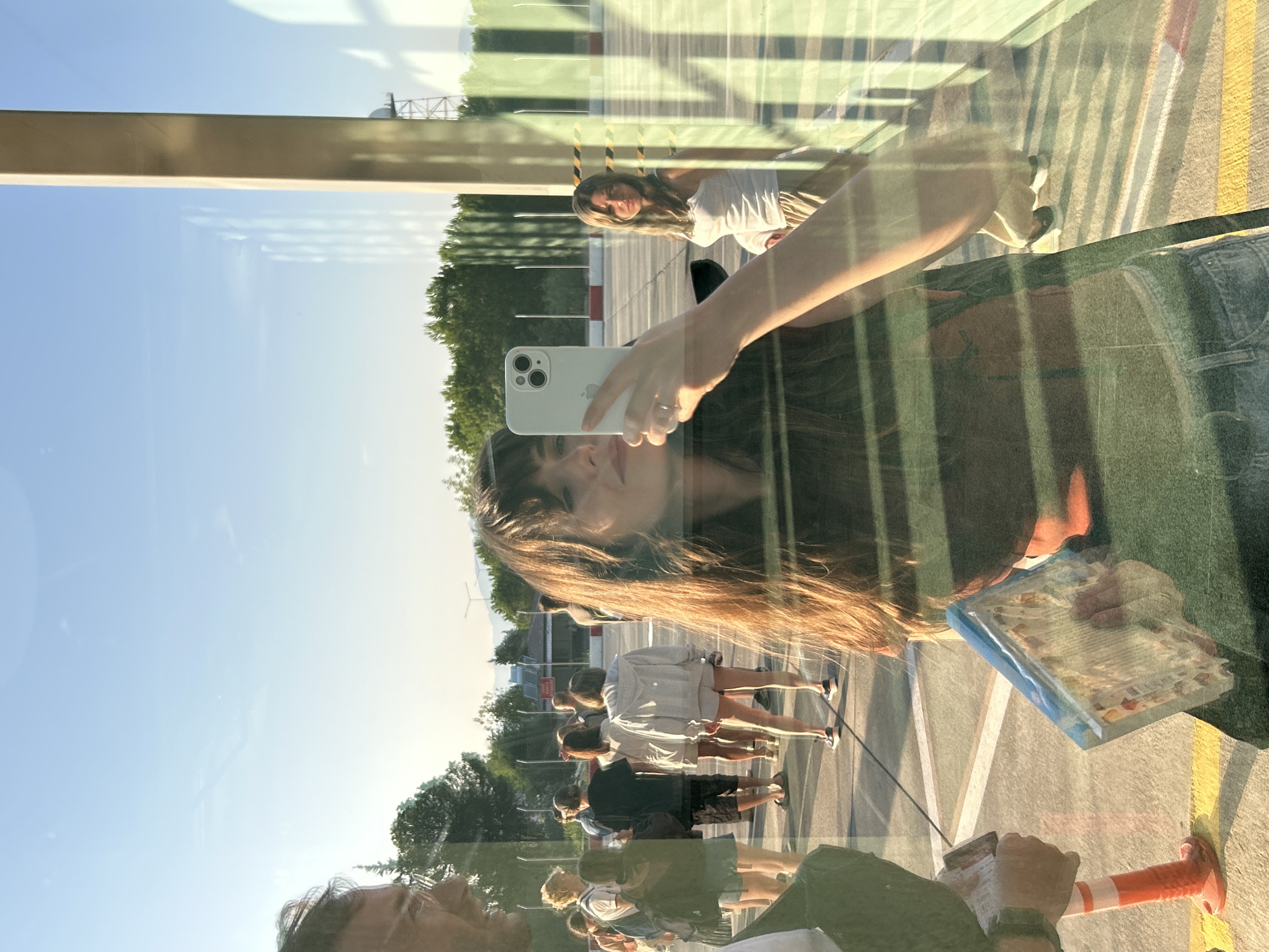 This is a selfie taken by a young woman with long, brown hair, who is holding an iPhone in her right hand. She is wearing a dark red top and denim shorts. Her left hand is visible, holding a book. She is reflected in a glass surface, likely a window, and behind her, a group of people are standing in what appears to be an outdoor area with a paved ground marked with yellow and white lines. The background shows trees, a fence, and a clear sky with some wispy clouds. A man with glasses and a green jacket is partially visible on the left, and another woman in a white tank top and light-colored pants is visible in the background to the right. The scene appears to be a public space, possibly an airport or a similar transit area, given the presence of a tower-like structure in the distance. The lighting suggests it is either early morning or late afternoon. The location is the city of Dalaman, Turkey.