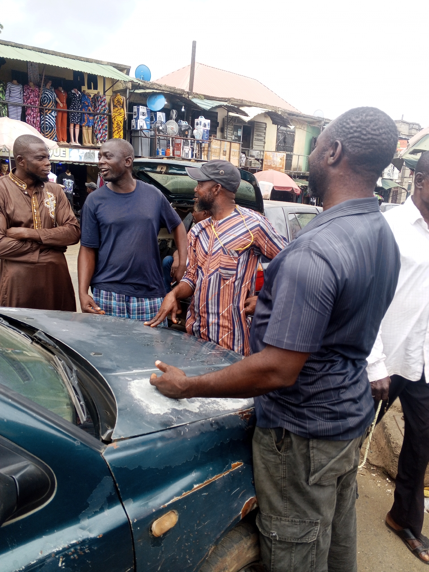 The image depicts a street scene in Okene, Nigeria, with several men gathered around a dark teal-colored car. The car appears to be old and shows signs of wear, with peeling paint and rust. The men are engaged in conversation, with one man in a brown robe with gold embroidery, another in a navy blue t-shirt and plaid shorts, a third in a striped shirt, and a fourth in a dark pinstripe shirt. A fifth man in a white shirt and black pants is partially visible on the right side of the image, holding a string of prayer beads. In the background, there are market stalls selling clothing and electronics. The sky is overcast.