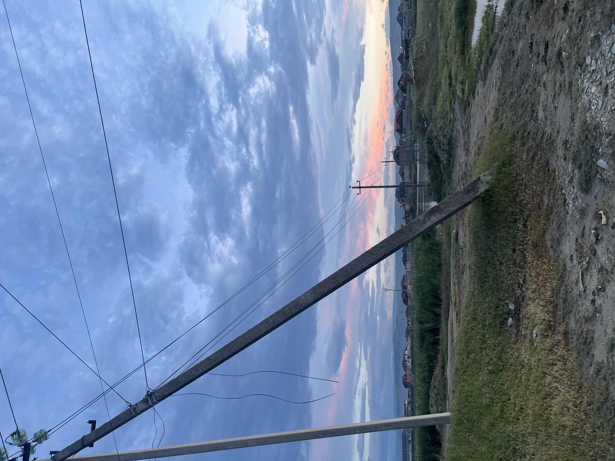 The photograph shows an outdoor scene in Kaspiysk, Russia, at what appears to be dusk or dawn, judging by the sky's colors. A concrete electrical or telephone pole is the prominent feature in the foreground, with wires extending across the frame towards the left and out of view. Some of the wires are broken and hanging down. A secondary metal support structure is visible below the concrete pole. In the background, houses are visible along the horizon line. The ground is a mix of grass and dirt, indicating a roadside or undeveloped area. The sky is filled with clouds, with colors ranging from blue to pink. There are no people visible in the scene.