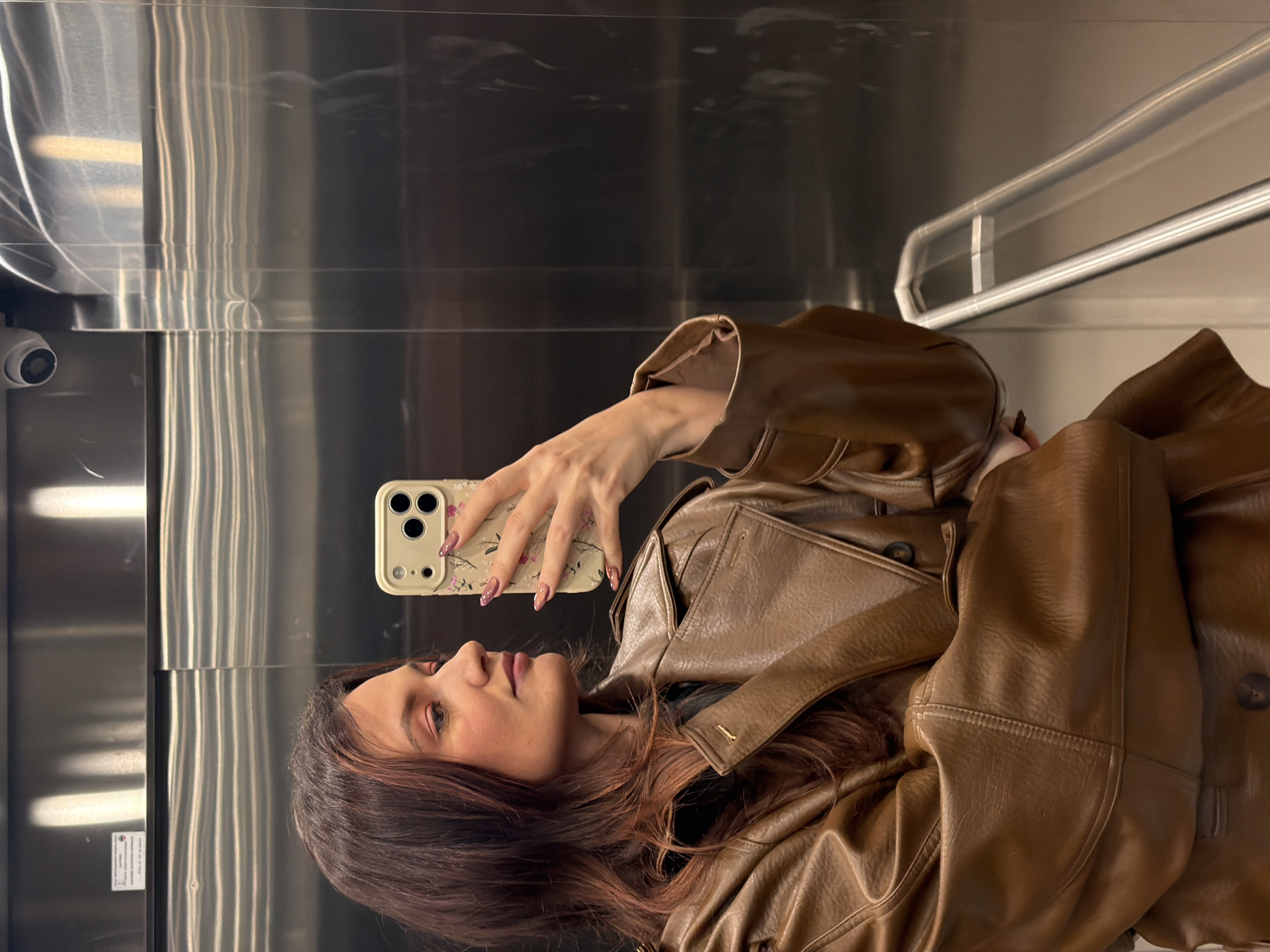 A young woman with long brown hair is taking a selfie in an elevator with stainless steel walls. She is wearing a brown leather jacket. Her phone has a beige case with a floral pattern. The elevator has a surveillance camera in the top left corner and a horizontal handrail in the upper right. The location is likely in Belgrade, Serbia.