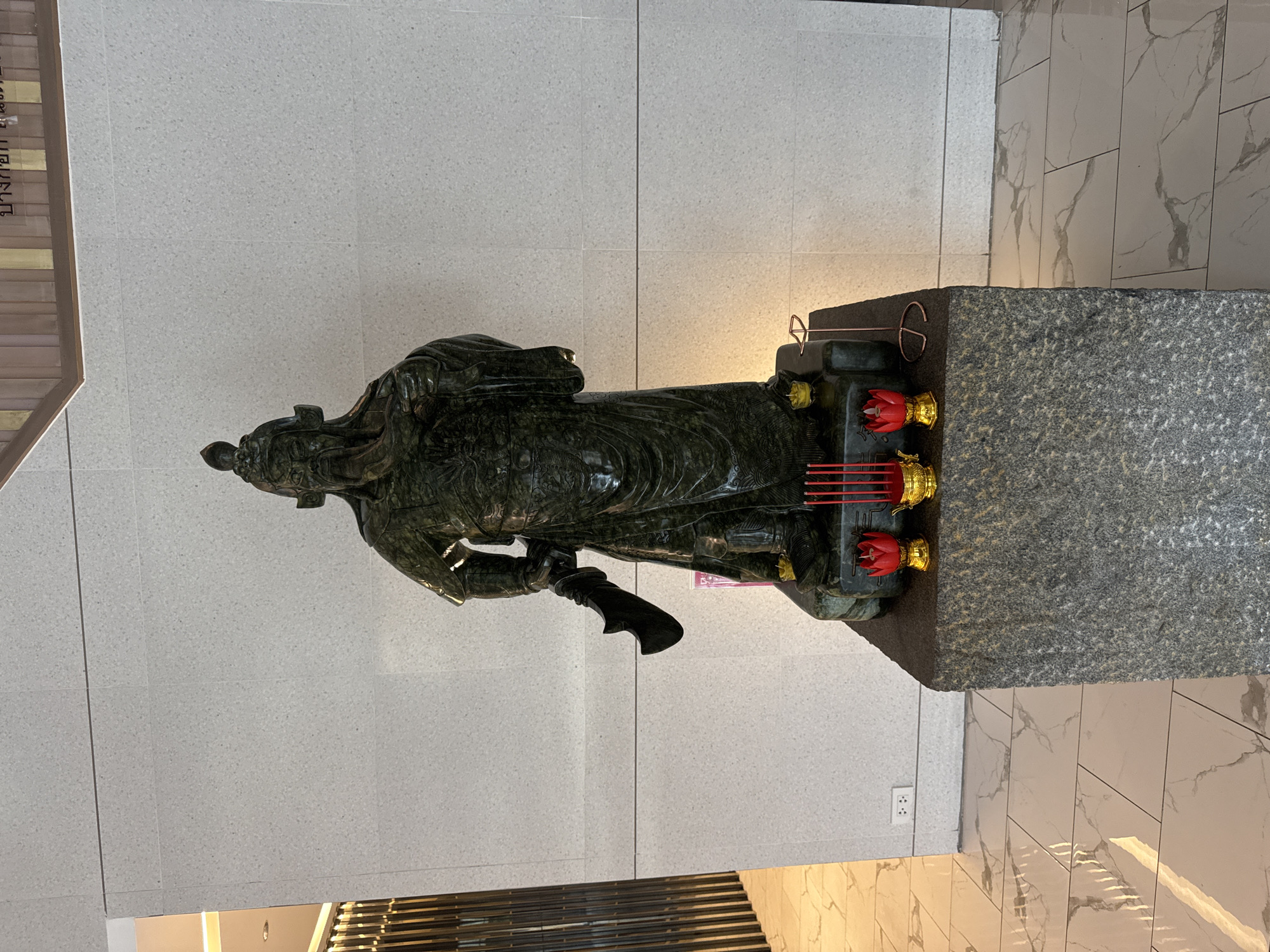 The image displays a large, dark green statue, likely carved from jade or a similar stone, depicting a revered figure in traditional attire. The statue is a male, possibly a warrior or deity, dressed in elaborate robes with detailed carvings and distinctive headwear. He holds what appears to be a weapon or staff in his right hand.
The statue is positioned on a sturdy, dark grey, rough-textured granite-like pedestal. On the pedestal in front of the figure, there are offerings: two red lotus-shaped candle holders with golden bases, and several thin red sticks, likely incense, placed in golden holders. A small, faded pink or purple slip of paper is tucked near the base of the statue.
The setting is indoors, with a light grey tiled wall featuring subtle speckles as the background. The floor is composed of white and grey marble-patterned tiles. Warm artificial lighting illuminates the statue and its immediate vicinity. An electrical outlet is visible on the wall. On the far left, a portion of a sign displays Thai text, with "บางกอก" (Bangkok) clearly legible, confirming the location context of Bangkok, Thailand. The overall scene suggests a place of cultural display or veneration.