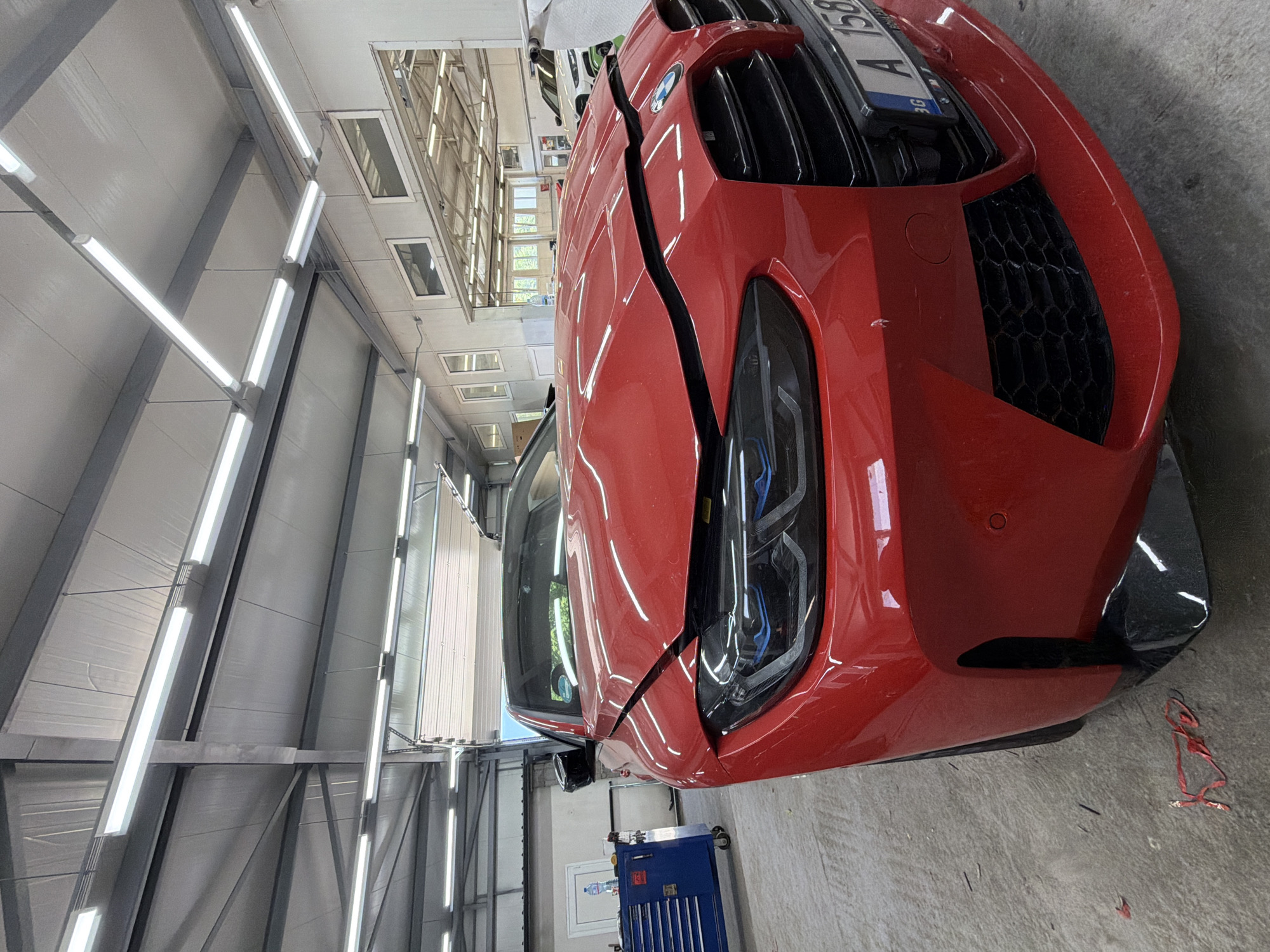 The image shows a red car, likely a BMW M3, parked indoors in what appears to be a garage or workshop. The car's front end is prominently displayed, with its distinctive grille and headlights visible. A long, dark crack runs down the center of the hood, suggesting some damage. The car is positioned at an angle, with the right side facing the viewer.
The setting is a well-lit interior space with white paneling on the walls and a concrete floor. Overhead, fluorescent lights illuminate the scene, and some industrial-looking metal beams and structures are visible. In the background, glimpses of other vehicles and what looks like a toolbox can be seen. A license plate with "A 158" is partially visible on the car's front. There is no indication of the time of day or weather conditions. The overall impression is of a car undergoing some kind of maintenance or repair in a professional setting. The location context of Burgas, Bulgaria is not evident from visual cues in the image itself.