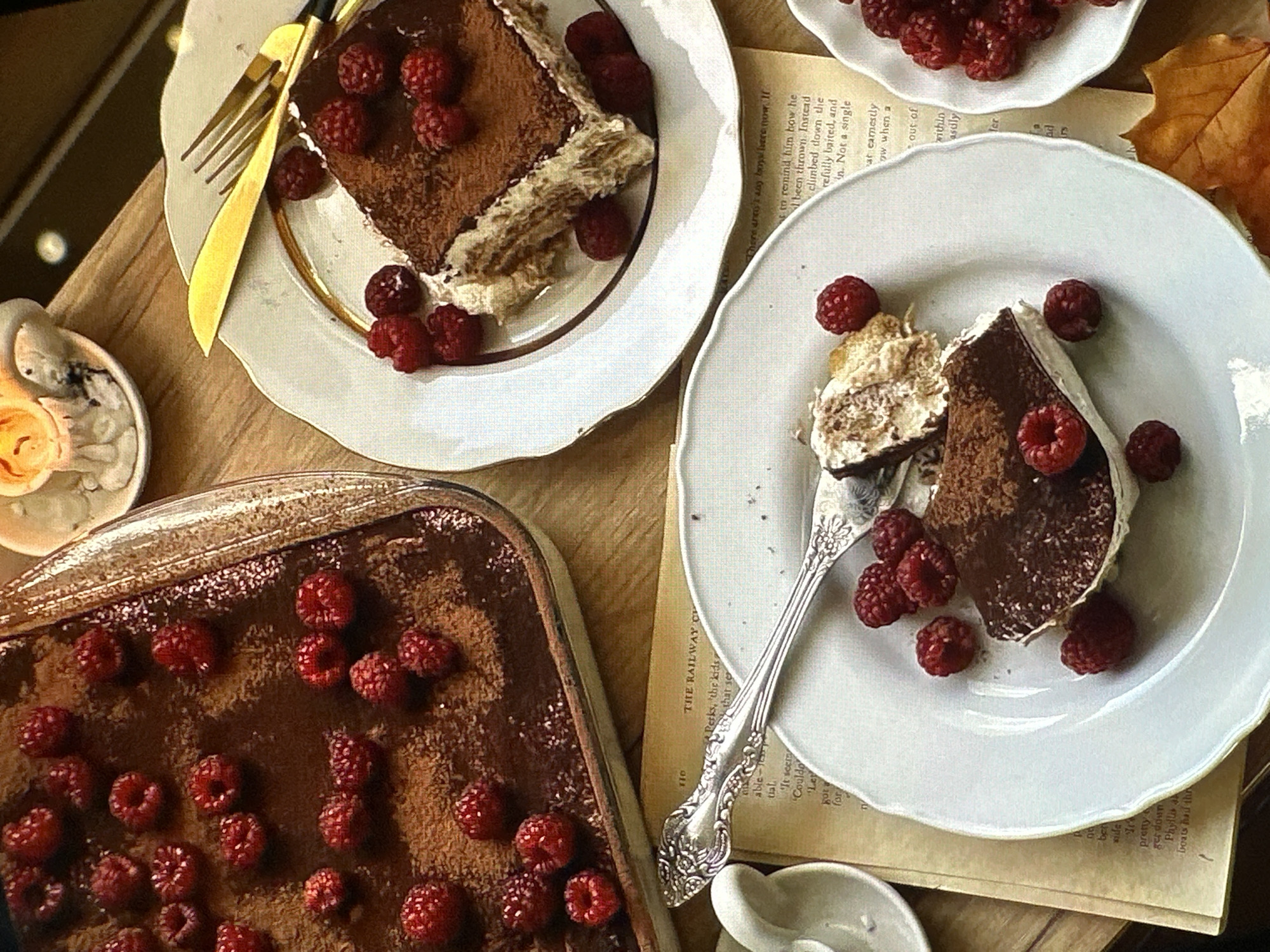A high-angle shot depicts a dessert setting. A glass baking dish filled with tiramisu topped with raspberries sits on a wooden surface, with a portion already cut out. Two additional slices of tiramisu are placed on ornate, white plates, each adorned with scattered raspberries. One plate has gold-colored cutlery beside it, while the other has a silver fork piercing a piece of tiramisu. A small, decorative cup, possibly a candle holder, is near the baking dish. A book with visible text lies under the plate with the silver fork. An autumn leaf is near the plate containing the raspberries. The scene is well-lit, creating a warm and inviting atmosphere. There is no indication of Yerevan, Armenia.