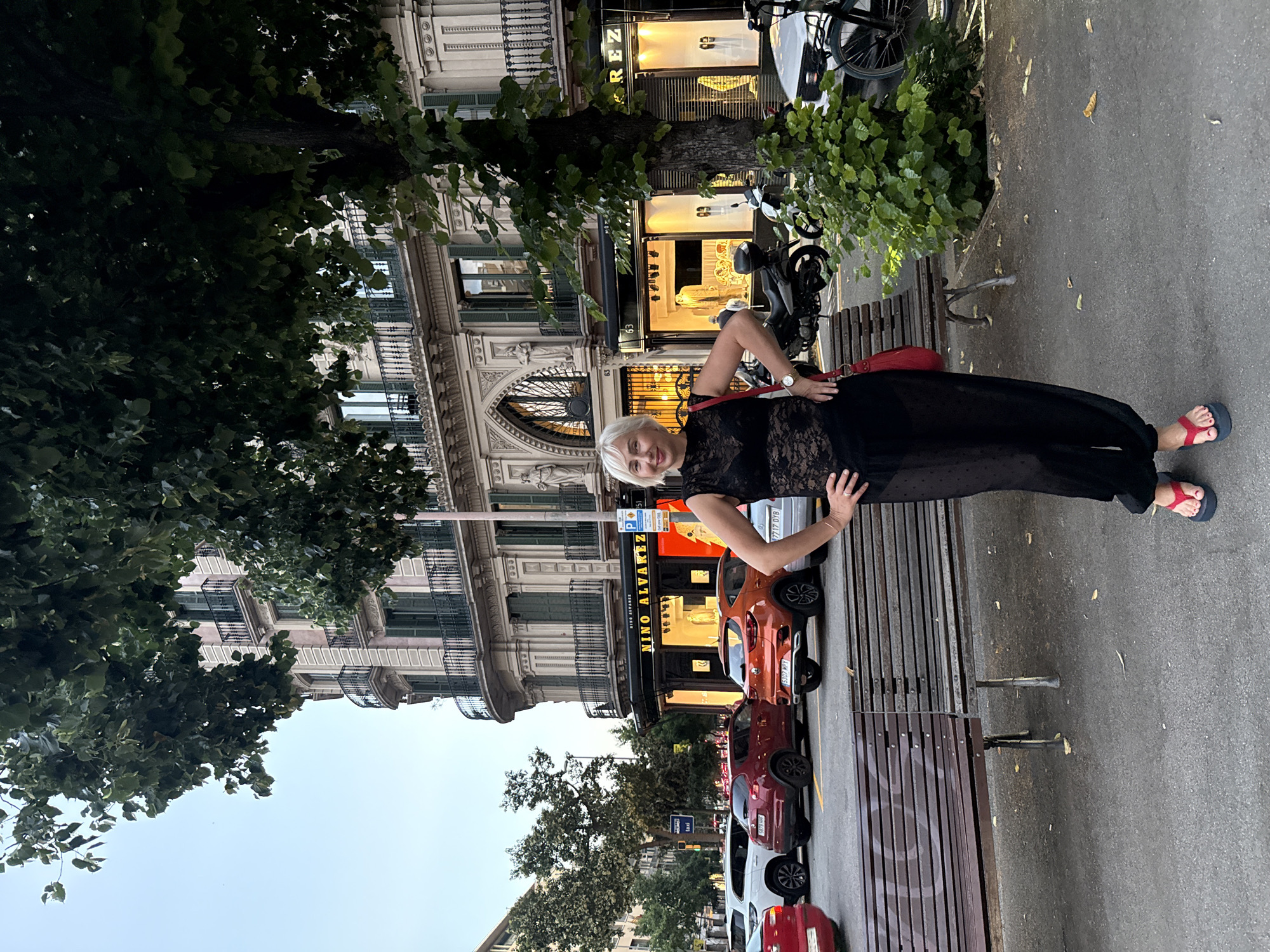 A Caucasian woman with short blonde hair and fair skin stands outdoors in front of a building. She is wearing a black lace top and black sheer pants, with red flip-flops. She has a red handbag on her left shoulder and her hands are on her hips. The building behind her is a multi-story structure with ornate architectural details, including balconies with black railings and decorative facades. A shop sign with "NINO ALVAREZ" is visible on the ground floor. Several cars are parked on the street in front of the building, including an orange SUV and a red car. A wooden bench is in the foreground, and a tree with green leaves is to the right of the woman, partially obscuring the building. The scene appears to be a street in Barcelona, Spain, likely during the day, given the lighting.