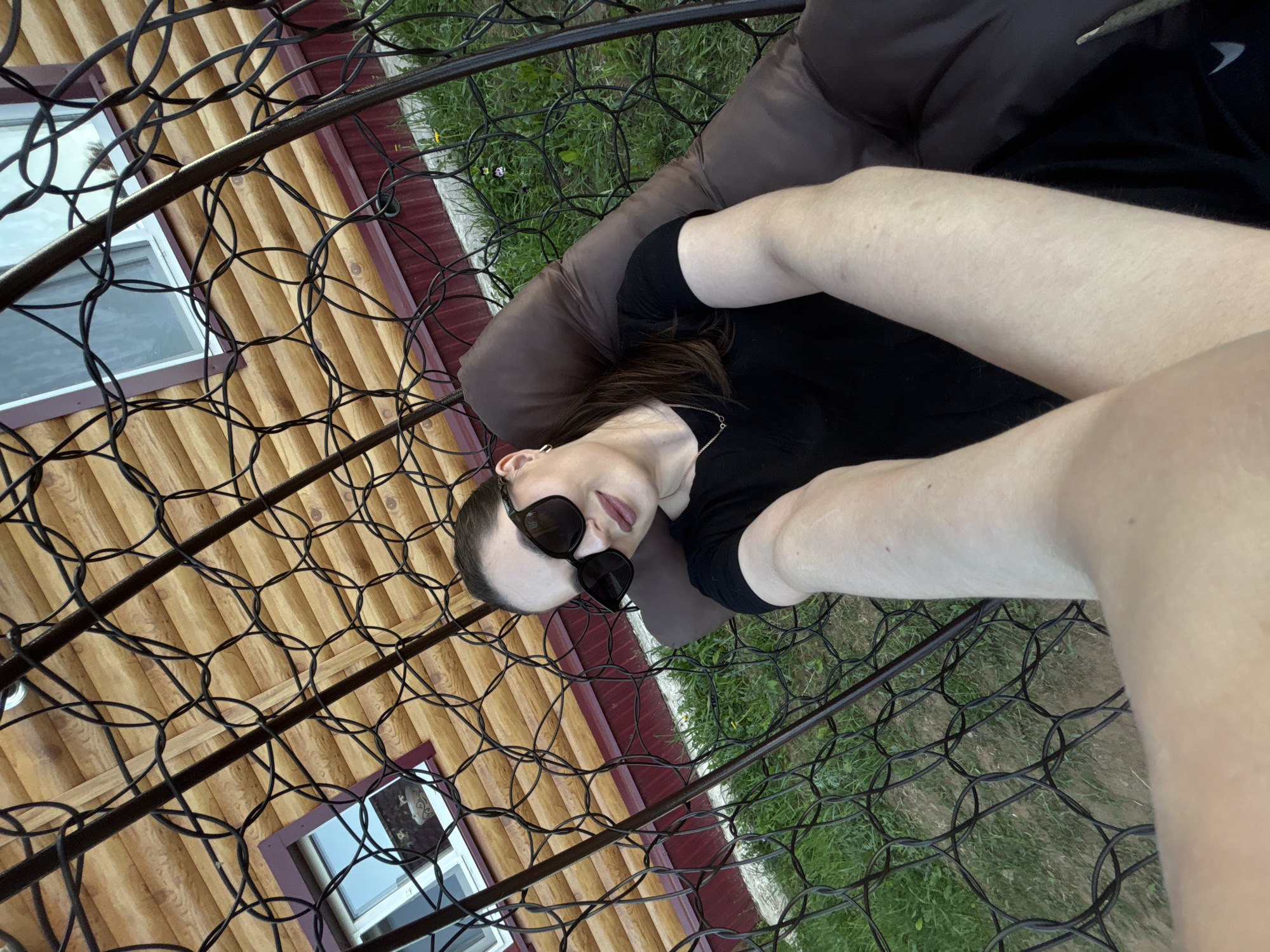 A woman wearing sunglasses and a black shirt is lying in a hanging chair or hammock. The structure of the chair is made of black metal and has a circular design. She is lying on a brown cushion. In the background is a log cabin with visible windows. Green grass is also visible outside the chair, next to the cabin. The photograph is taken from an elevated angle, looking down towards the subject and surroundings. The location cannot be determined from the image.