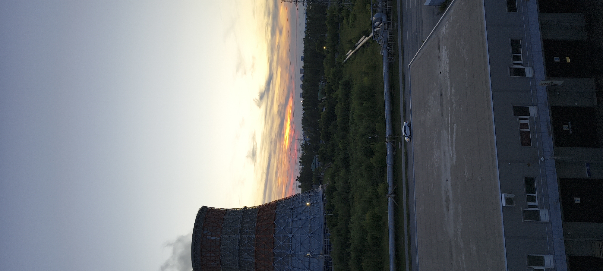 The image shows an industrial landscape at dusk. On the left, a large cooling tower emits steam into the pale sky. The tower is a cylindrical structure with a red and grey patterned exterior. In the foreground, a grey, flat-roofed industrial building with multiple loading bays is visible. A white car is parked on the road in front of this building. Behind the cooling tower and to the right, a dense area of green trees stretches across the middle ground. Beyond the trees, pipes and power lines are visible, along with some distant buildings and a utility tower. The sky displays a gradient of colors from pale blue at the top to warm oranges and yellows near the horizon, indicating the setting sun. The overall scene depicts an industrial area under a twilight sky.
