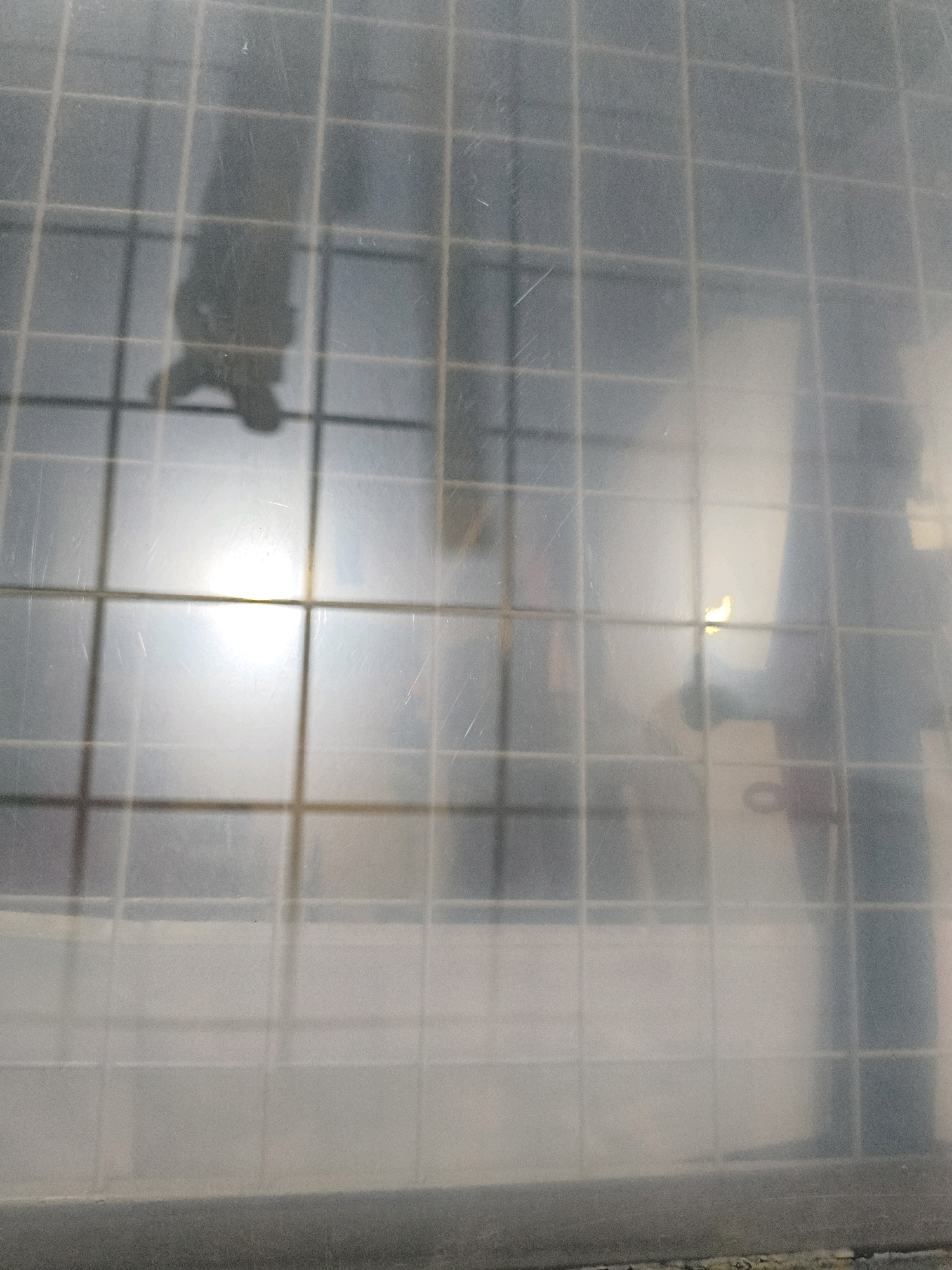 The image depicts a blurry, gridded surface, possibly a window or partition, with indistinct figures and light sources visible behind it. The overall impression is of a diffused, indoor environment. The grid pattern suggests a structured setting. One figure appears to be standing, with another possibly hanging upside down, and there is a bright light source to the left. The environment is somewhat hazy, obscuring finer details. The location is stated to be in Sukasejati, Indonesia, though the image itself does not contain any direct visual cues to confirm this.