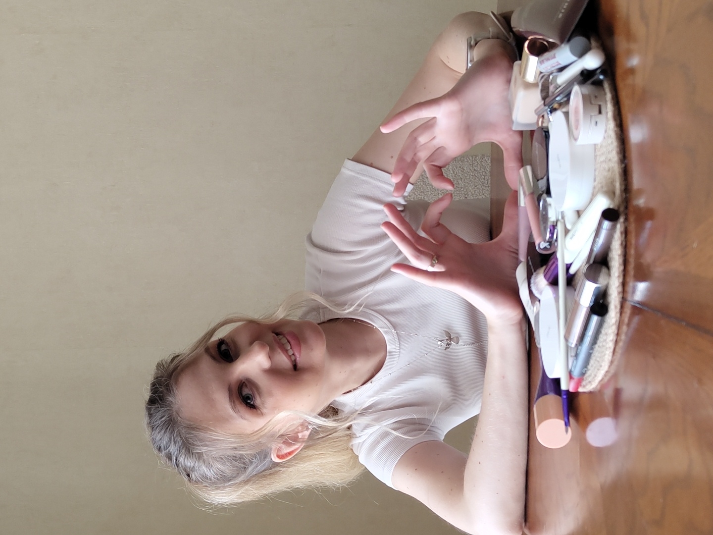 A young Caucasian woman with blonde hair pulled into a ponytail and wearing a white t-shirt is seated at a wooden table. She is making a heart shape with her hands. In front of her on the table is a collection of makeup products, including brushes, lipsticks, and compacts, arranged on a circular mat. The background is a plain, light-colored wall. The image does not contain any identifiable landmarks or context that would place it in Krasnodar, Russia.