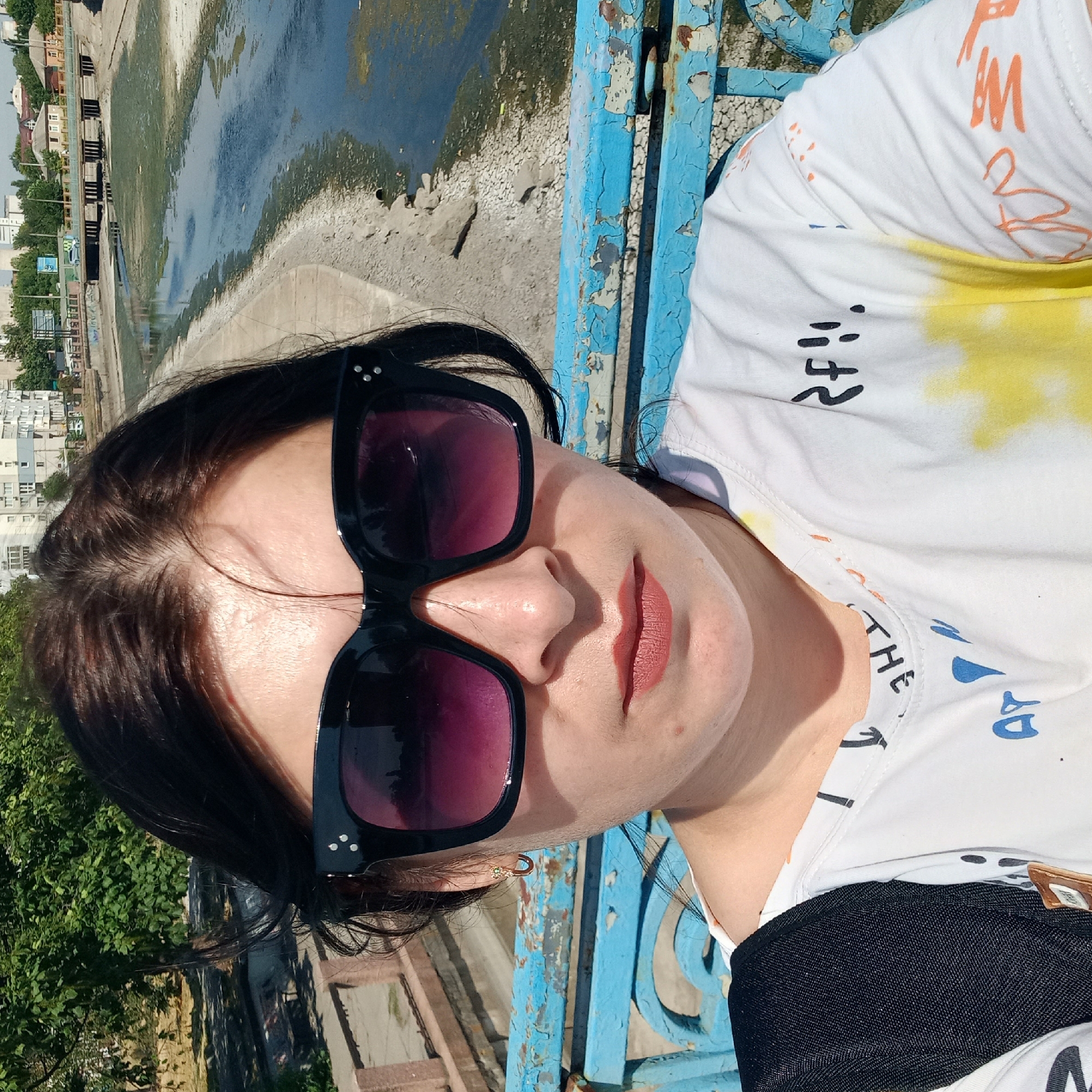 The image is a close-up selfie of a woman wearing large black sunglasses and a white t-shirt with a colorful tie-dye pattern and black and blue lettering. She is standing on a bridge with a blue, weathered railing. In the background, a dried-up riverbed with some water is visible, along with a concrete embankment and buildings in the distance. The scene appears to be in Kropyvnytskyi, Ukraine, as indicated by the characteristic urban architecture and the presence of a river channel. The woman's hair is dark and pulled back, with some strands falling across her face. She has a gold earring with a small green stone in her left ear. The overall lighting suggests it is daytime, likely sunny.
