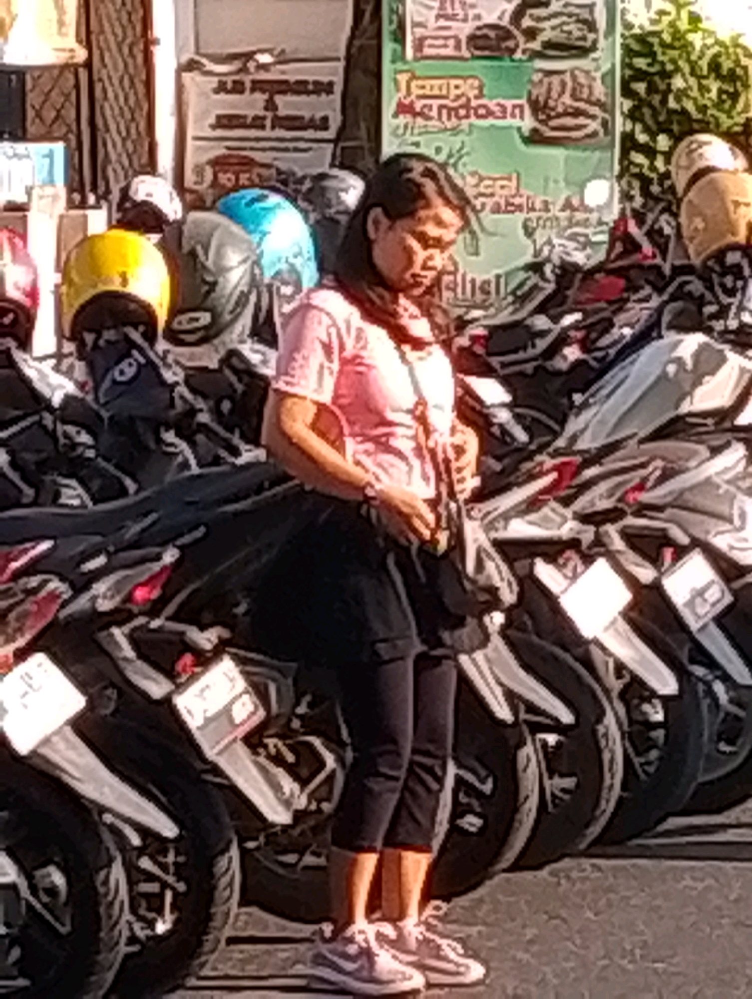 The image captures a street scene in Probolinggo, Indonesia, likely in the morning or late afternoon given the lighting. A woman wearing a pink t-shirt and black pants stands amidst a row of parked motorcycles. Her gaze is directed downwards, suggesting she might be tending to the vehicles or interacting with someone off-camera. Behind her, a collection of helmets in various colors, including red, yellow, blue, and green, are visible, indicating a potential motorcycle rental or service point. The background features a building with signage that appears to be in the local language, hinting at commercial activity. The overall atmosphere is that of a busy urban environment with a focus on motorcycle transportation.