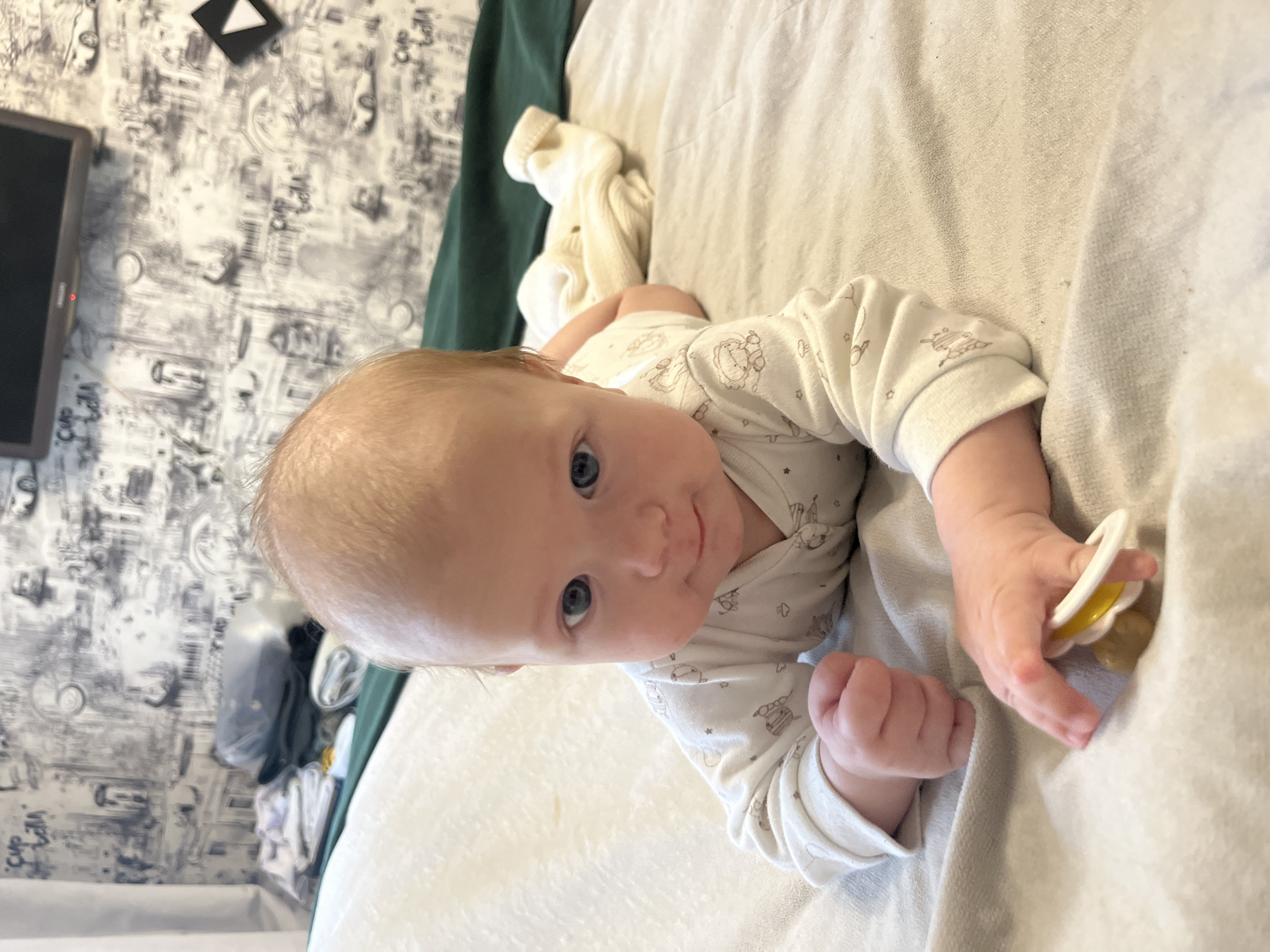 The image shows a baby lying on its stomach on a bed. The baby is wearing a white onesie with a pattern of animals. The baby is looking at the camera with its blue eyes. The baby's hands are near its face, and it is holding a pacifier. The background of the image is a bed with a light gray duvet and a dark green blanket. The wall behind the bed is covered with wallpaper that has a black and white pattern of buildings and cars. There is a television on the wall above the bed. The scene is indoors and appears to be in a bedroom. It is not possible to determine the location in the city of Andreyevo, Russia based on the provided image.