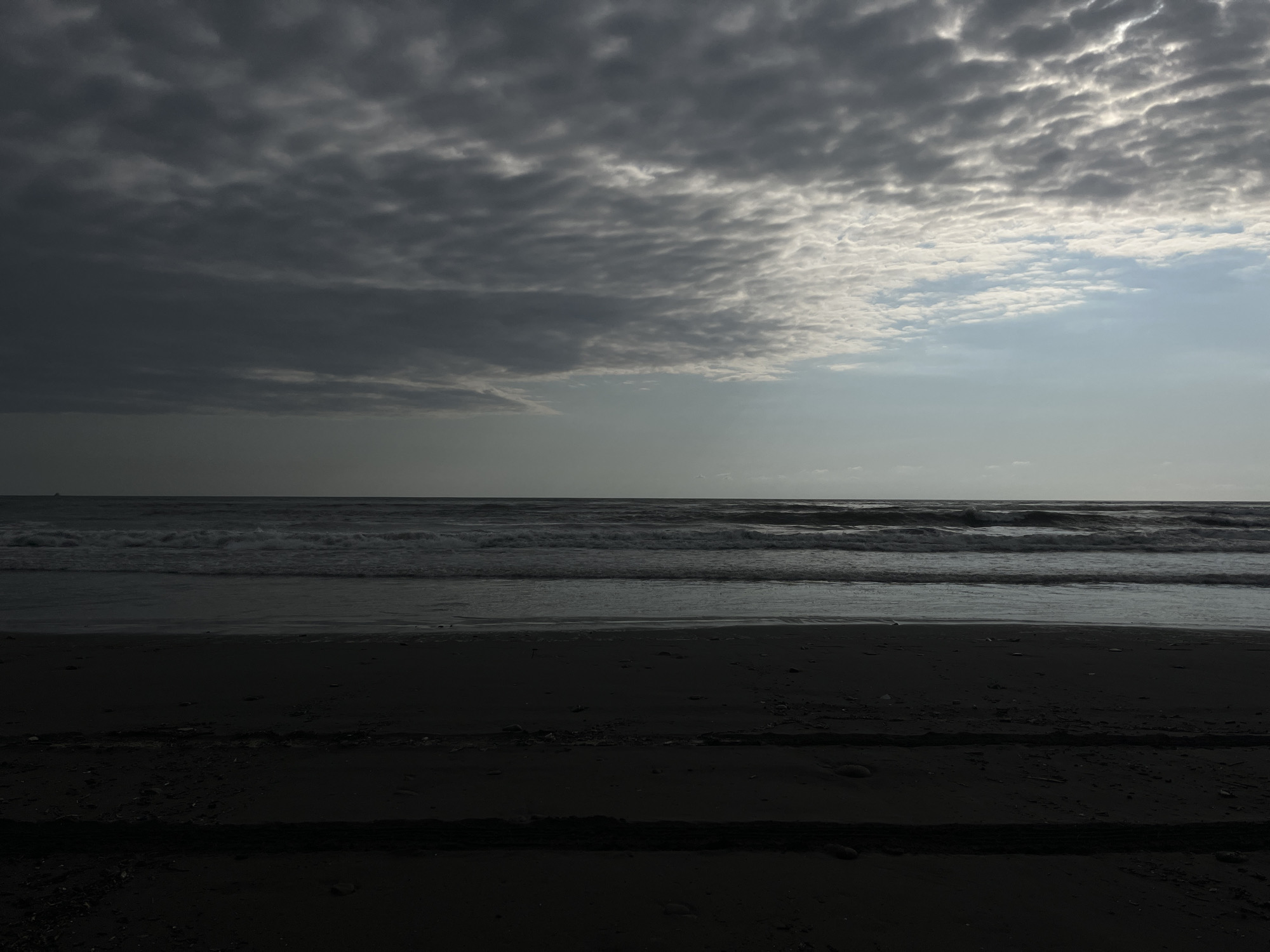The image depicts a dark, overcast beach scene, likely in the city of Supsa, Georgia, based on the provided context. The foreground is dominated by dark, wet sand, with faint tire tracks or indentations visible. Beyond the sand, the ocean stretches out, with moderate waves breaking on the shore, creating white foam. The water is a dark, choppy gray. The horizon line is distinct, separating the sea from the sky. The sky itself is heavily clouded, with darker, heavier clouds on the left side of the frame and lighter, brighter clouds on the right, where the sun is partially breaking through, casting a pale light. There are no people or distinct structures visible in the scene. The overall atmosphere is moody and somber, suggesting either early morning, late evening, or impending bad weather.