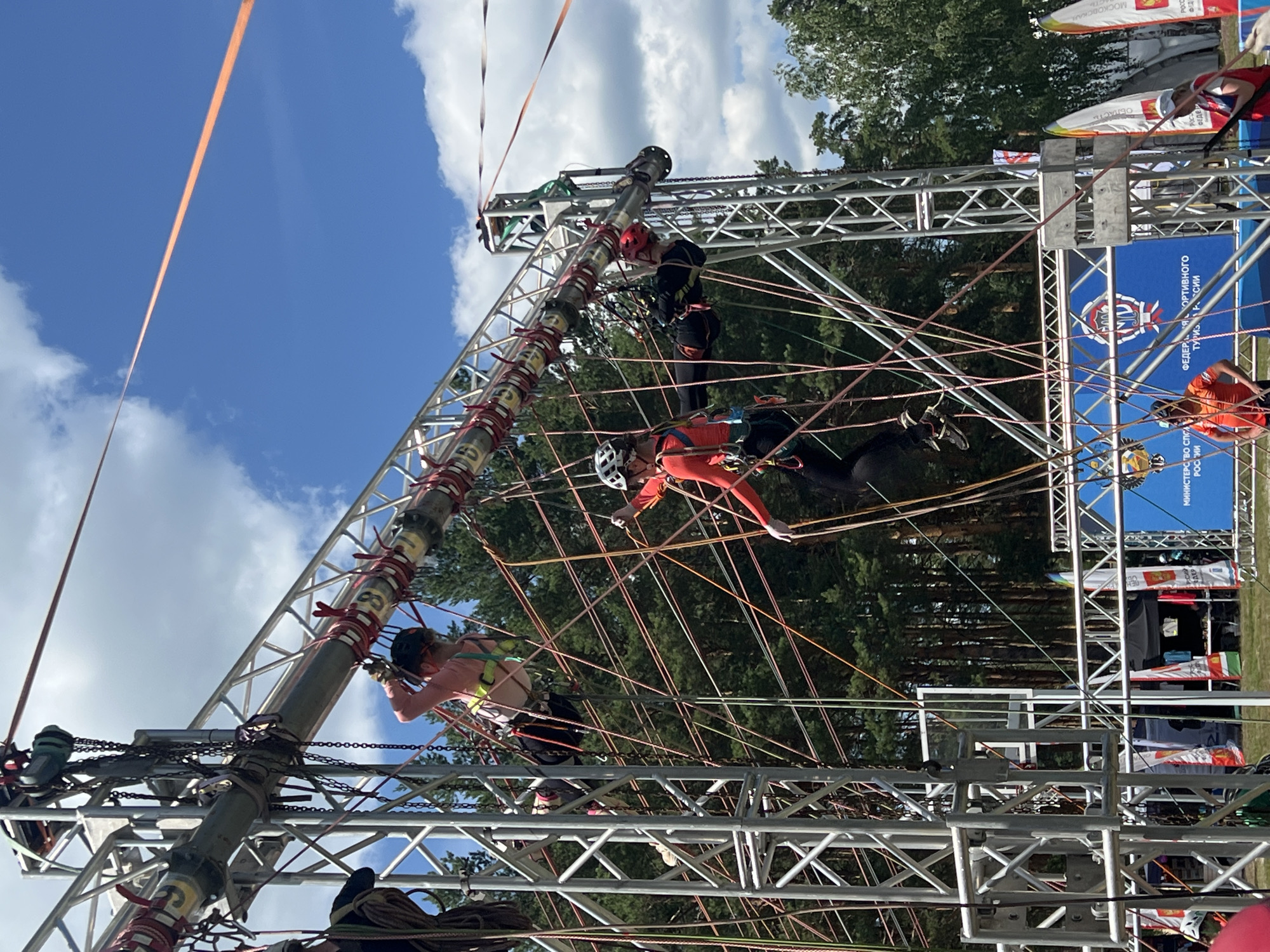 In Penza, Russia, on a bright, cloudy day, an outdoor obstacle course is set up. The course consists of a large metal truss structure with numerous ropes and climbing apparatuses suspended from it. Several individuals, dressed in athletic wear and helmets, are actively participating in the course, navigating the ropes and challenges. One person in a pink top and black pants is suspended mid-air, engaged with the ropes. Another individual in an orange long-sleeved shirt and black pants is also navigating the obstacle. In the background, more participants can be seen, along with tents, banners, and a densely wooded area. The overall scene depicts a sporting event or training exercise taking place outdoors.