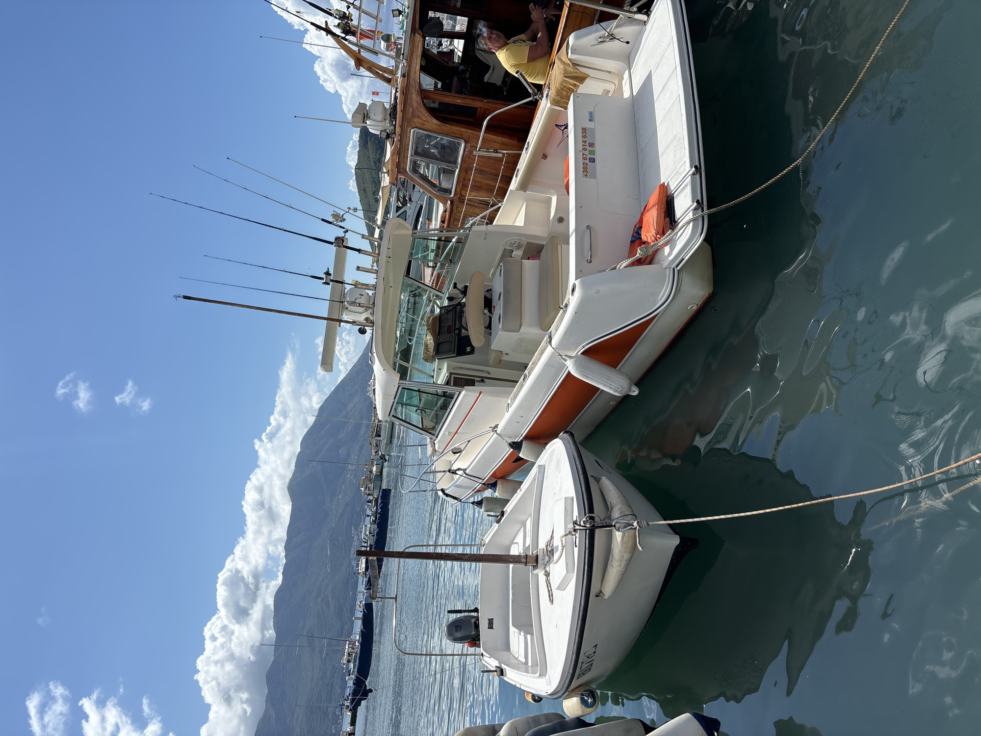 The image depicts a sun-drenched marina scene in Budva, Montenegro, with several boats moored in calm, greenish-blue water against a dramatic mountainous backdrop. In the midground, a white pleasure boat with orange trim is prominently displayed, equipped with multiple fishing rods in rod holders along its side. An orange life vest is visible, and a white sticker on its hull features social media icons (Instagram, Facebook, YouTube, TikTok, WhatsApp) and a phone number: "+382 67 44 638".
To its immediate right, a smaller white dinghy with an outboard motor and several fenders is also docked, with a wooden pole affixed to its bow. Behind the white pleasure boat, a larger, traditional wooden cabin cruiser is moored. A person wearing a yellow shirt is seated inside its cabin, appearing relaxed and looking towards the camera, with what looks like smoke or vapor being exhaled.
The background features a majestic mountain range partially covered by large, white cumulus clouds, under a clear blue sky. More boats are visible further along the marina. The bright daylight and serene water suggest a pleasant day, likely midday or early afternoon, with an atmosphere of leisure and coastal activity.